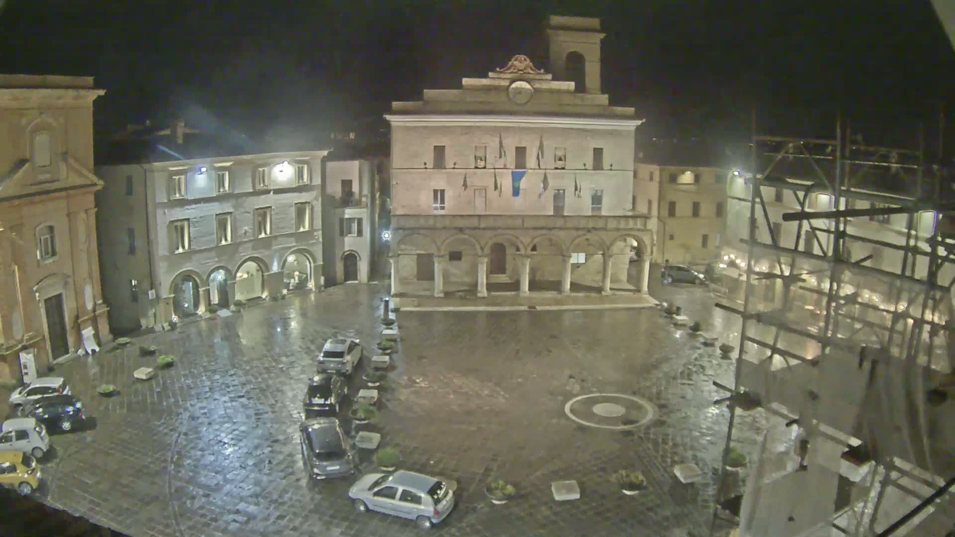 A rain-slicked town square at night, centered on a large building with arched entryways, surrounded by other buildings and a few parked cars.