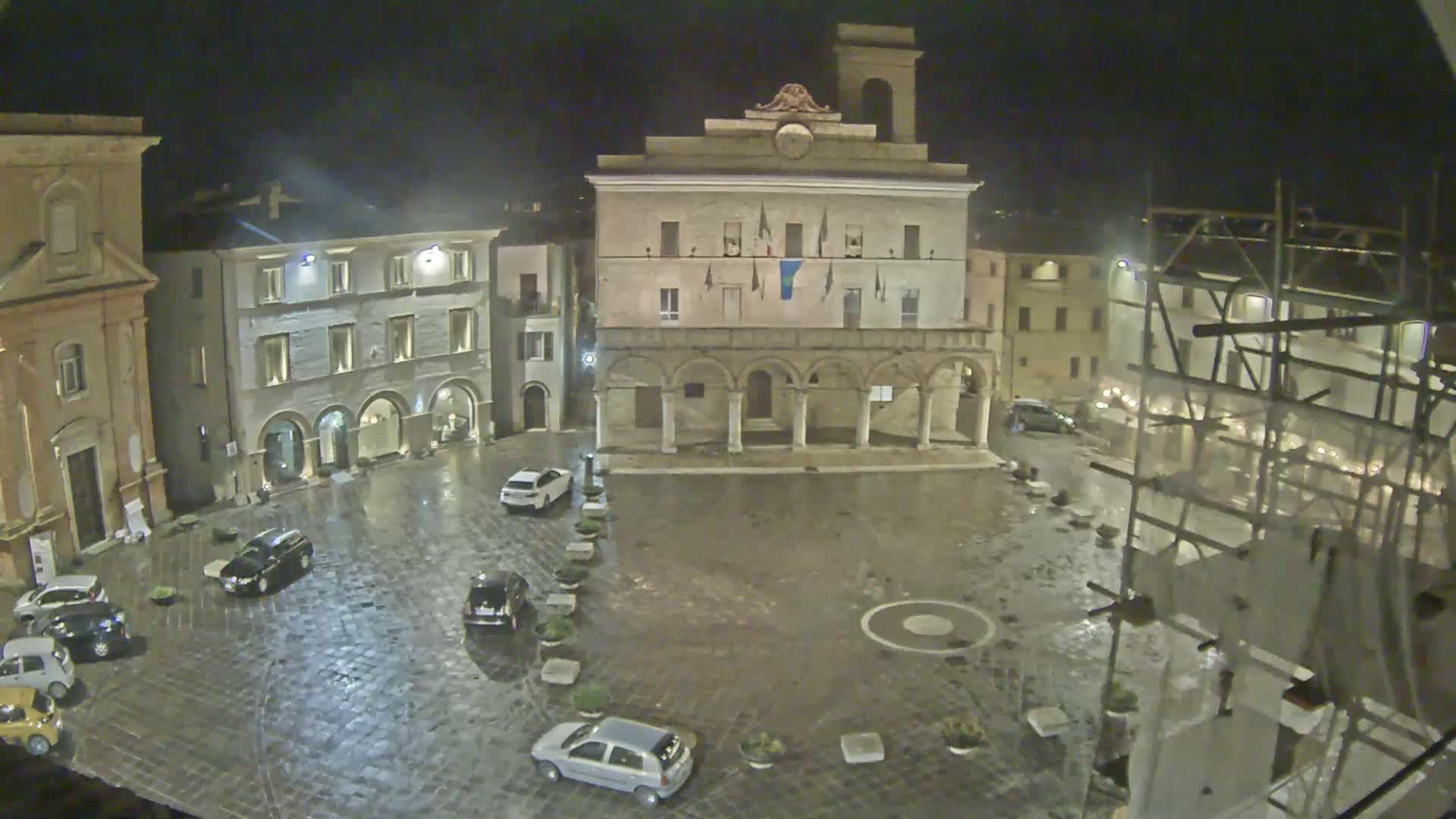 A rain-slicked town square at night, centered around a large, arched building with several cars parked around its perimeter, under scaffolding on the right.