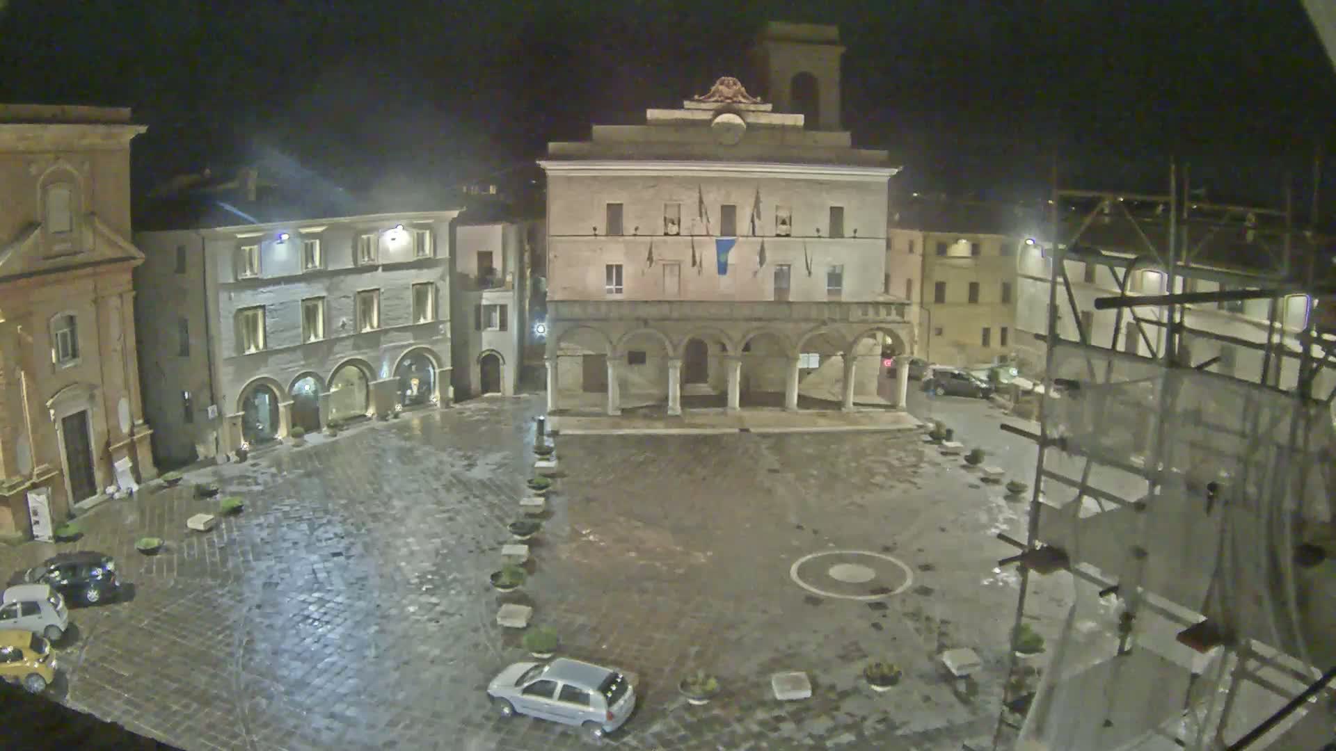 A rain-slicked cobblestone town square at night is centered around a large, arcaded building, with several smaller buildings and a few cars visible.