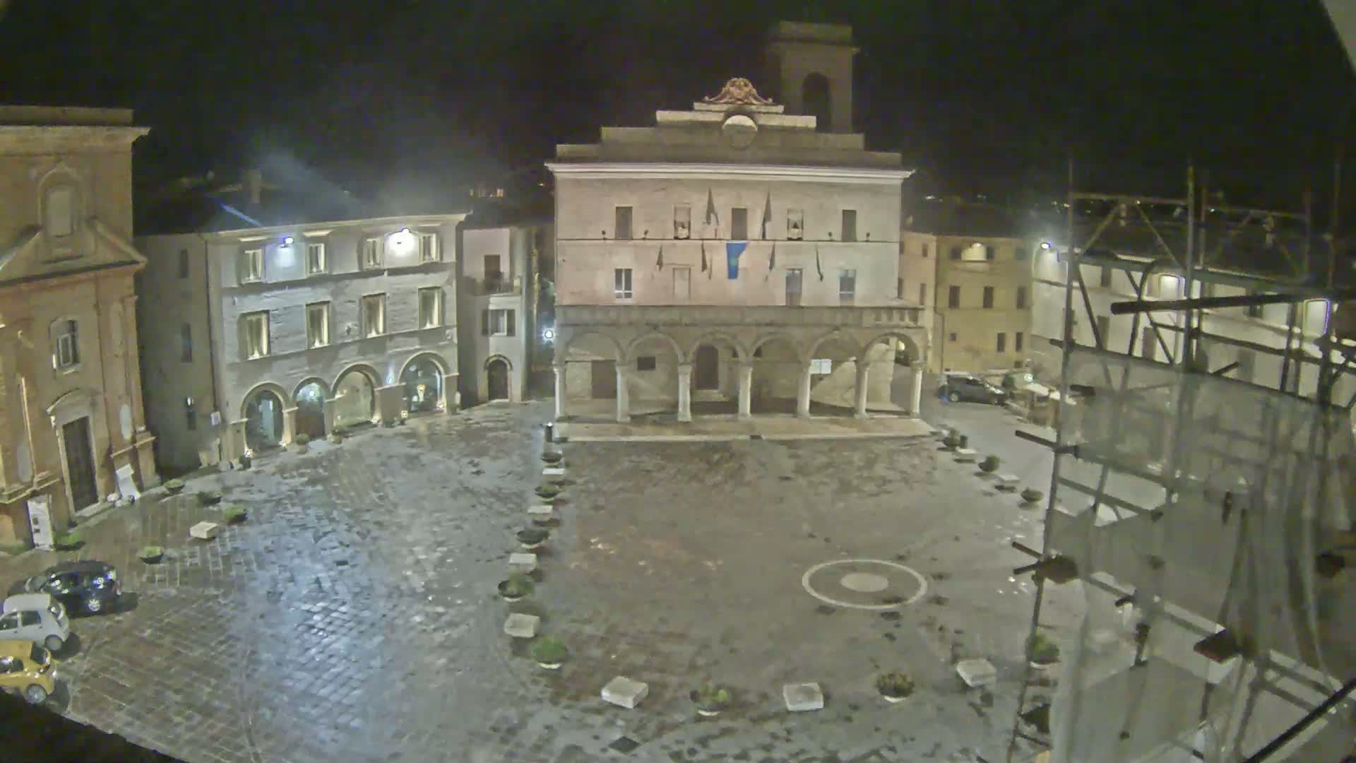 A wet, nighttime view of a town square dominated by a large, arched building, surrounded by other buildings and a few parked cars.
