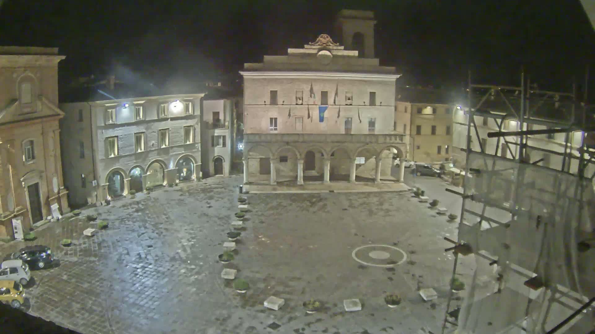 A wet, cobblestone town square at night is centered around a large, arched building with several smaller buildings surrounding it.