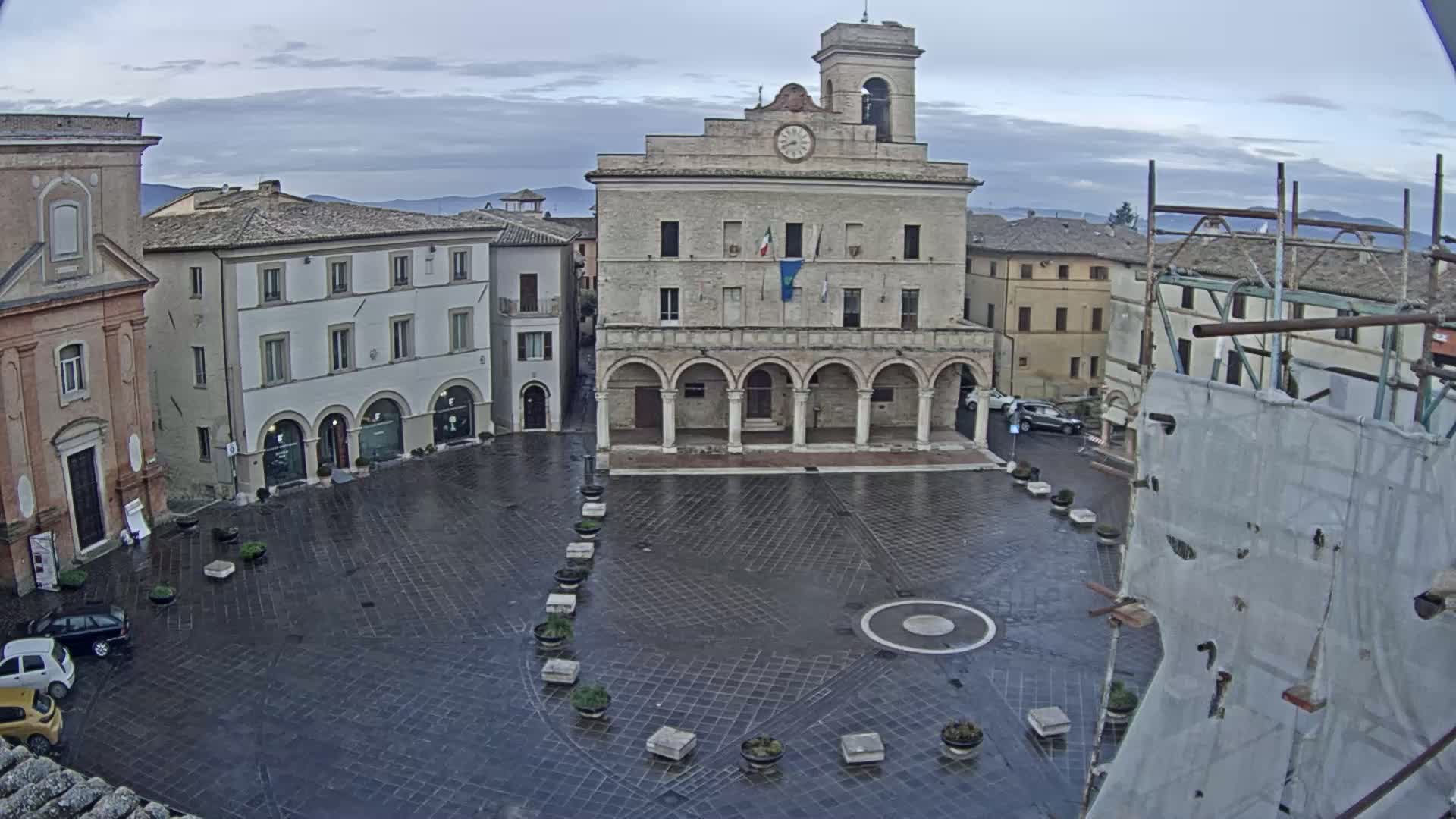 A town square, wet from recent rain, is centered on a large, classical-style building with arched entryways under an overcast sky.