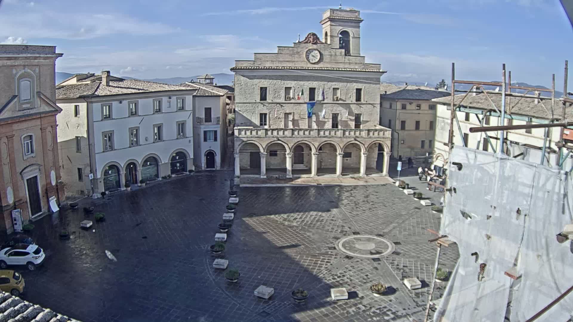 A mostly empty town square, under a partly sunny sky, is centered on a large, light-colored building with arched ground-level entrances.