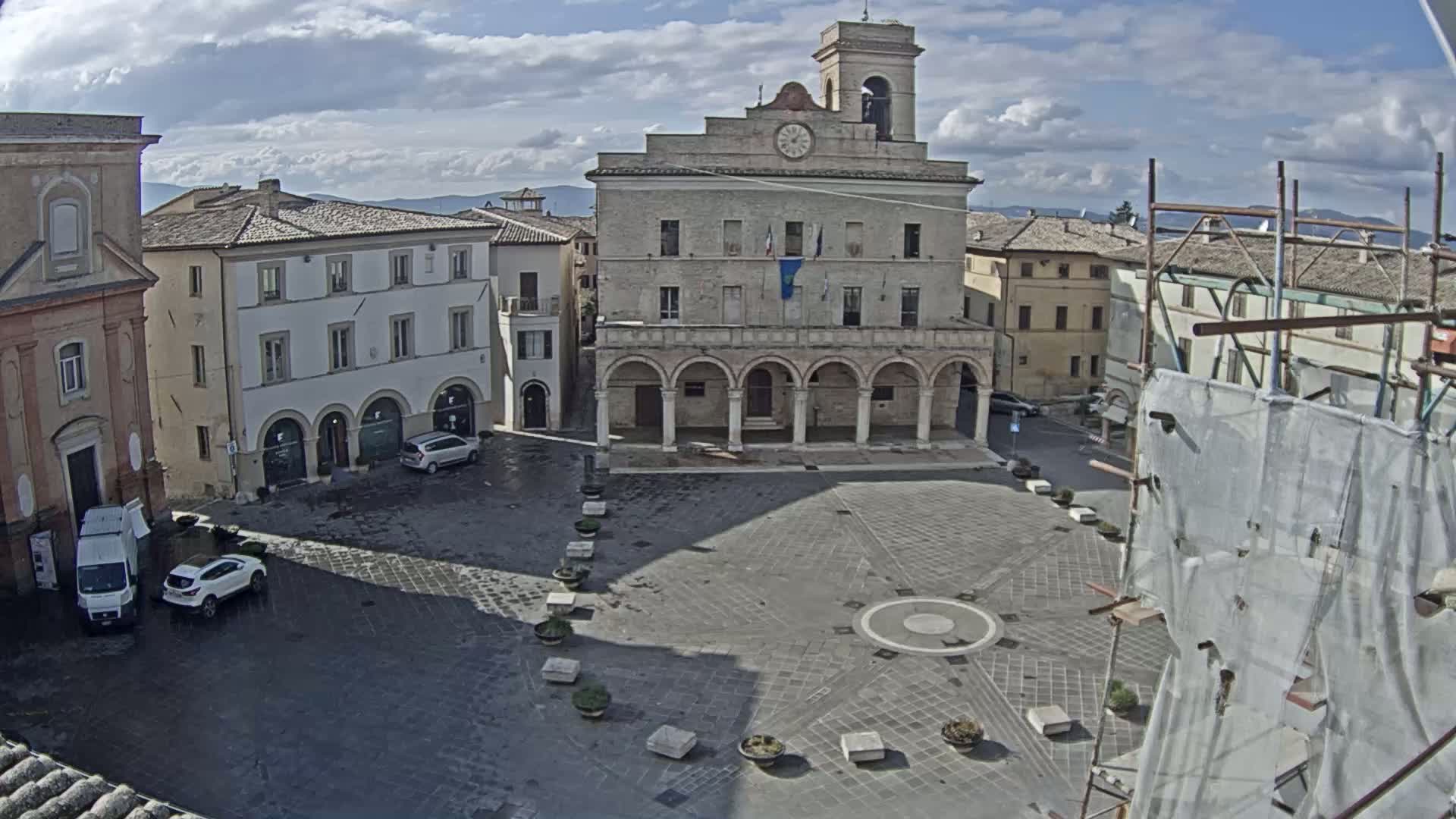 A large town square with a stone building featuring arched colonnades, surrounded by other buildings, under a partly cloudy sky.