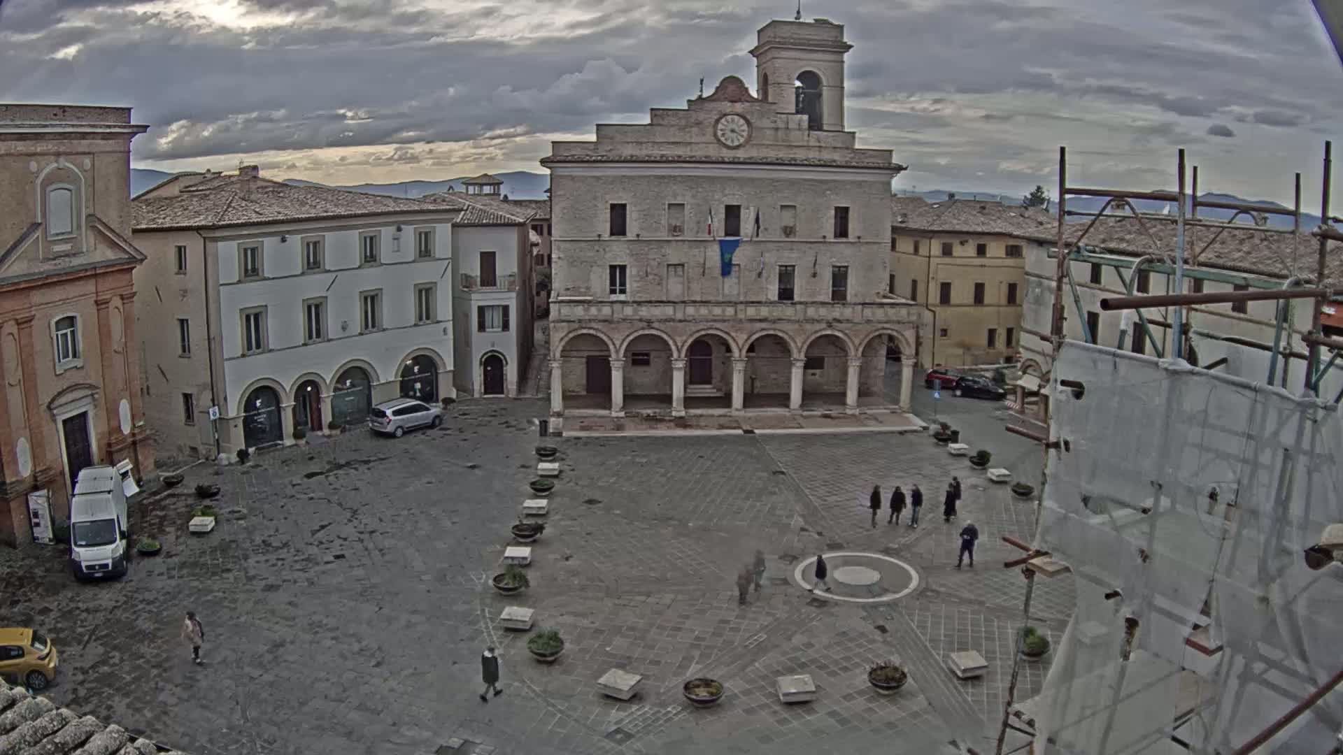 An overcast day reveals a large town square with a stone building featuring arched entryways and a clock tower, surrounded by other buildings and a few pedestrians.