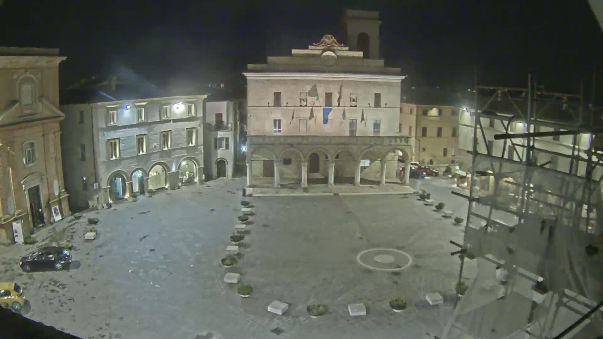 An empty town square at night, illuminated by artificial light, features a large building with arches on one side, and scaffolding on another.