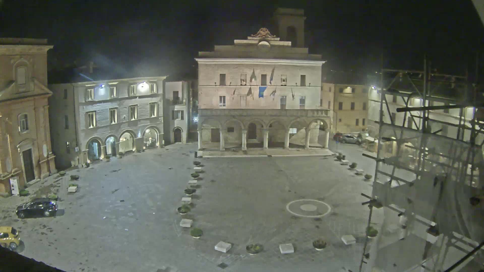 A nighttime view of a town square with several buildings, including a large stone structure with arches, under a dark, possibly cloudy sky.