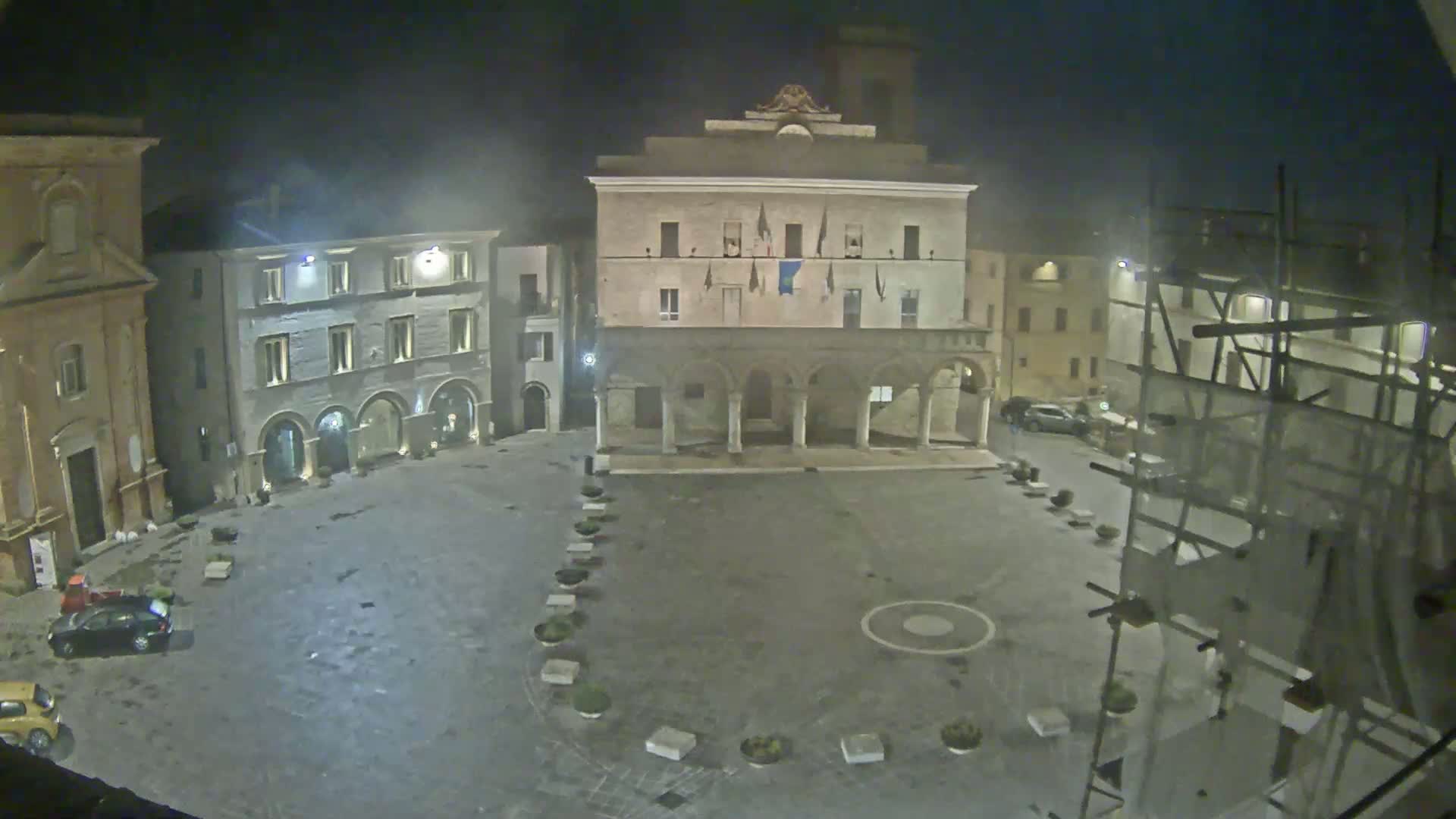 A mostly empty town square at night, under a hazy sky, is surrounded by historic buildings, one of which is under construction.