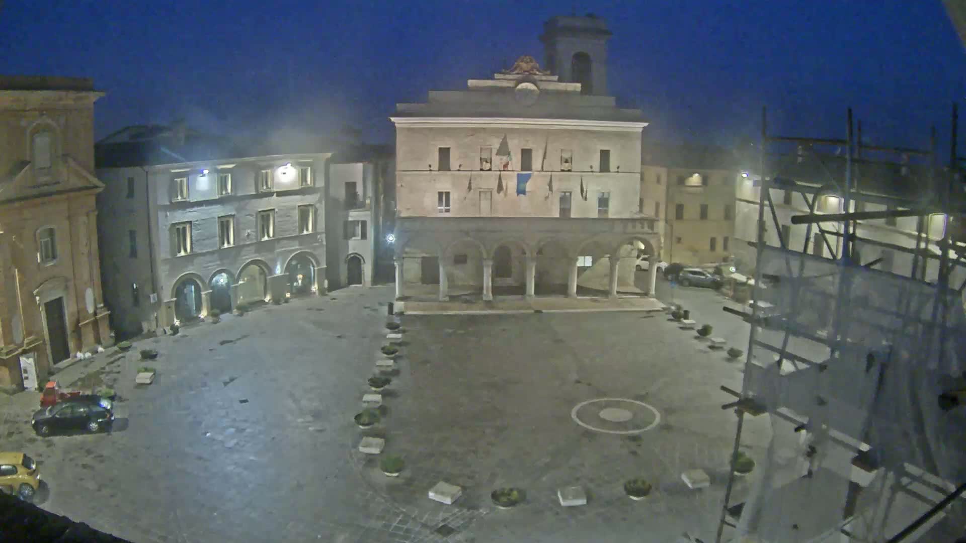 An empty town square at night, under a dark sky, is surrounded by historic buildings, one of which is a large, arched structure.