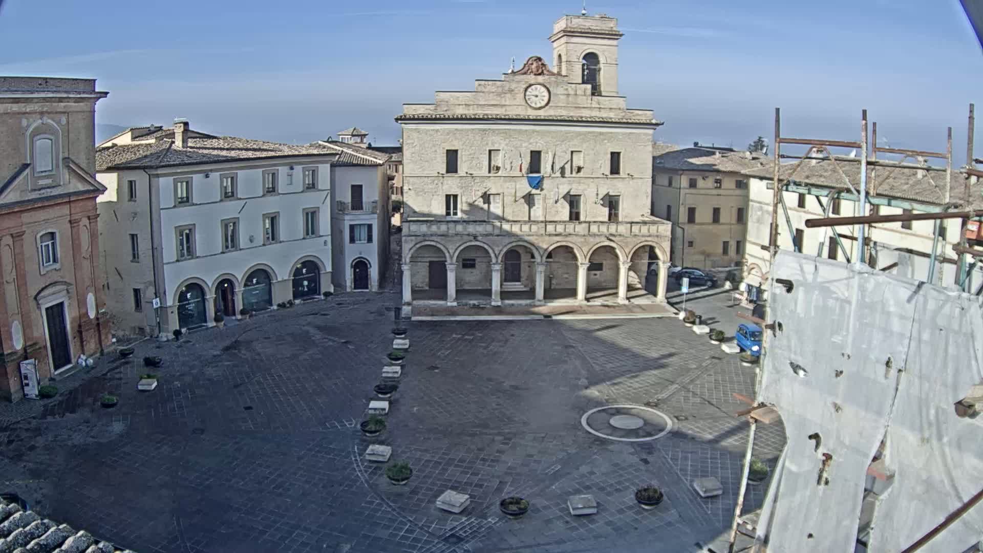 A large, empty town square is centered on a stone building with arched entryways, surrounded by other buildings under a clear, sunny sky.