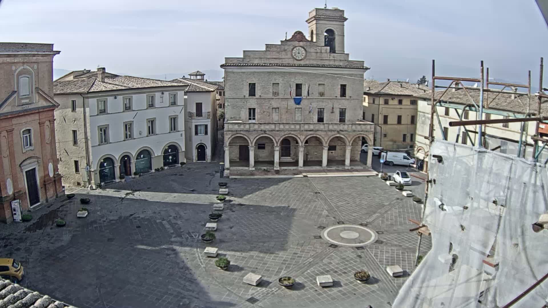 A town square with a large stone building, surrounded by other buildings, under a mostly sunny sky.