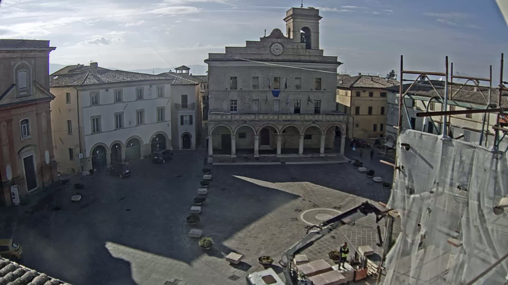A town square with a large, stone building featuring arched walkways, surrounded by other buildings under a mostly sunny sky.