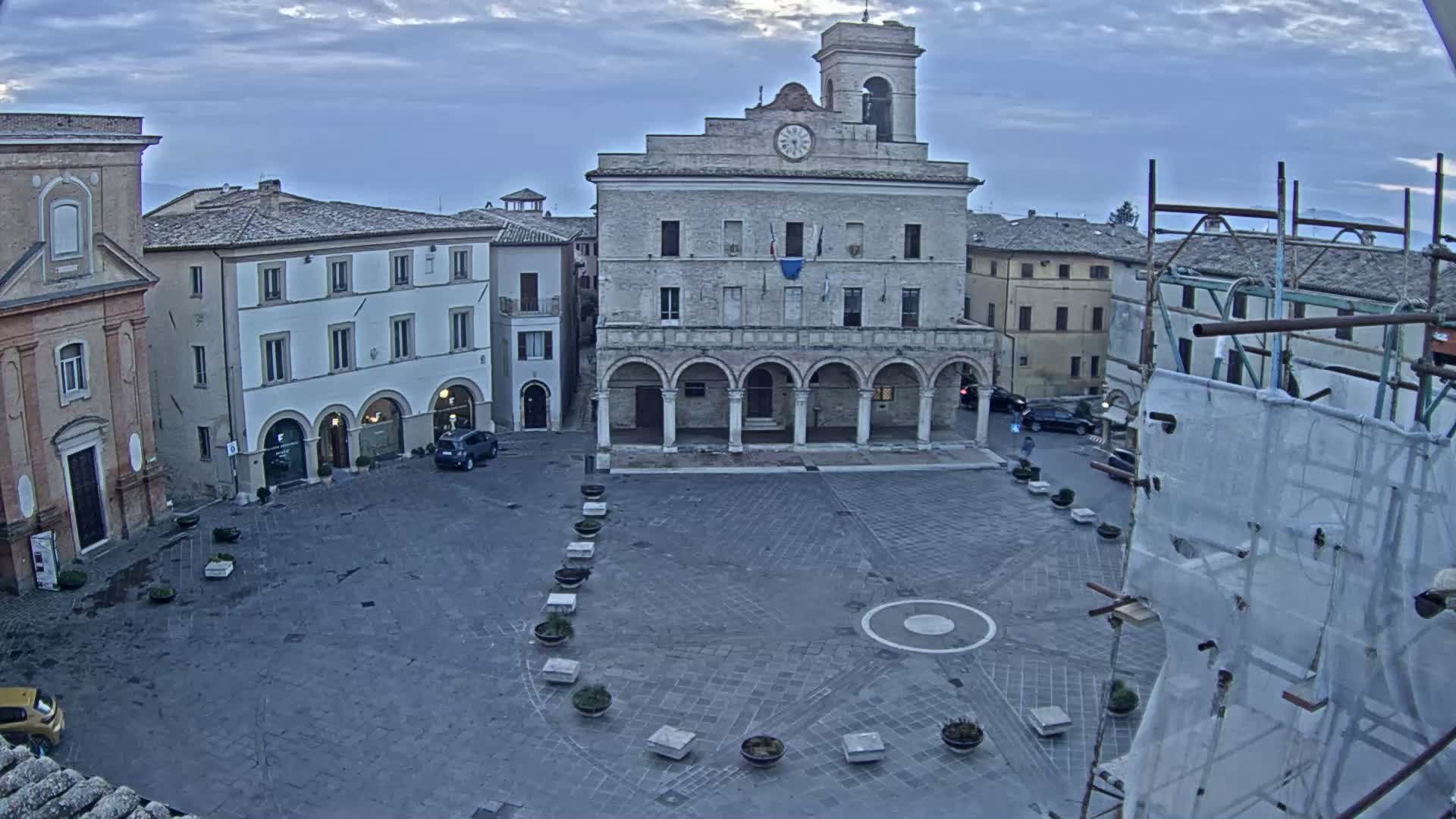 A large town square under an overcast sky is centered around a large, pale stone building with arched entryways.