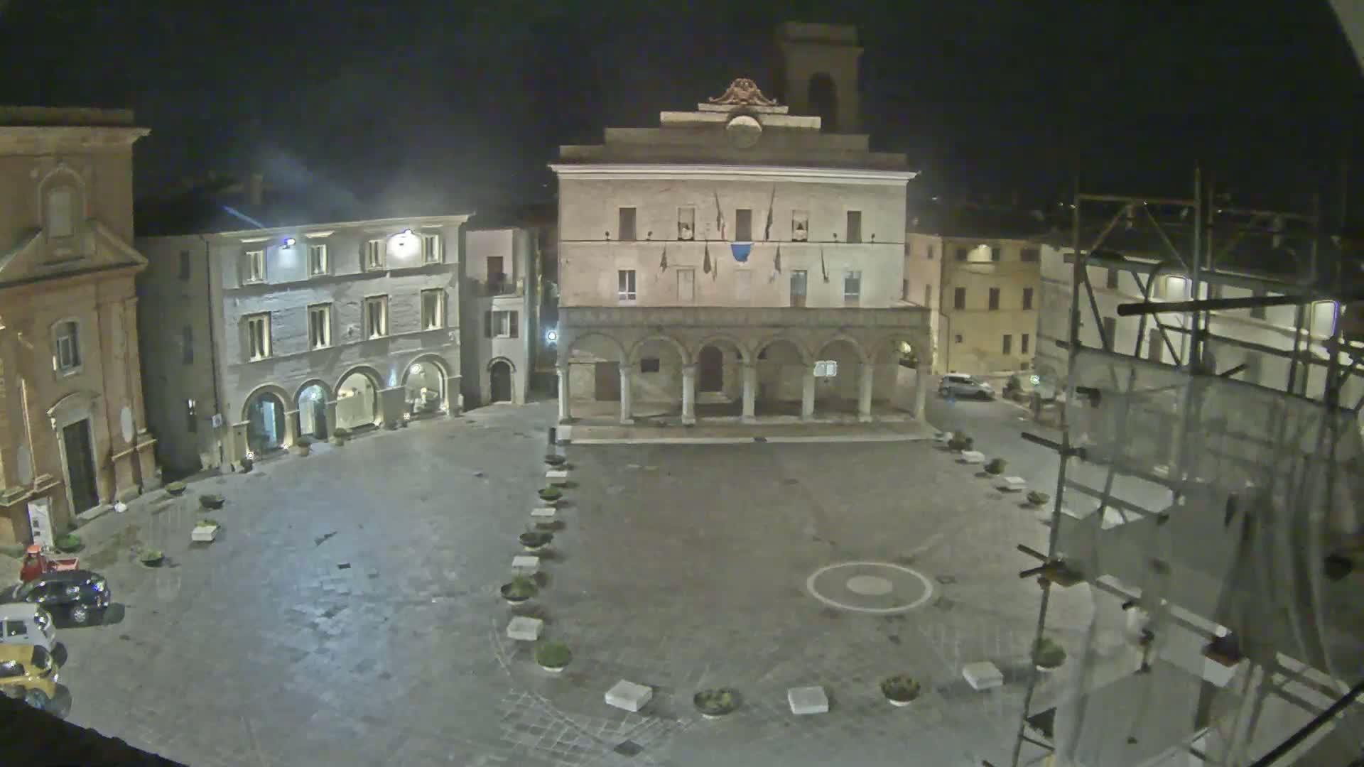 A nighttime view of a town square, featuring several buildings, including a large structure with arches, under a dark sky.