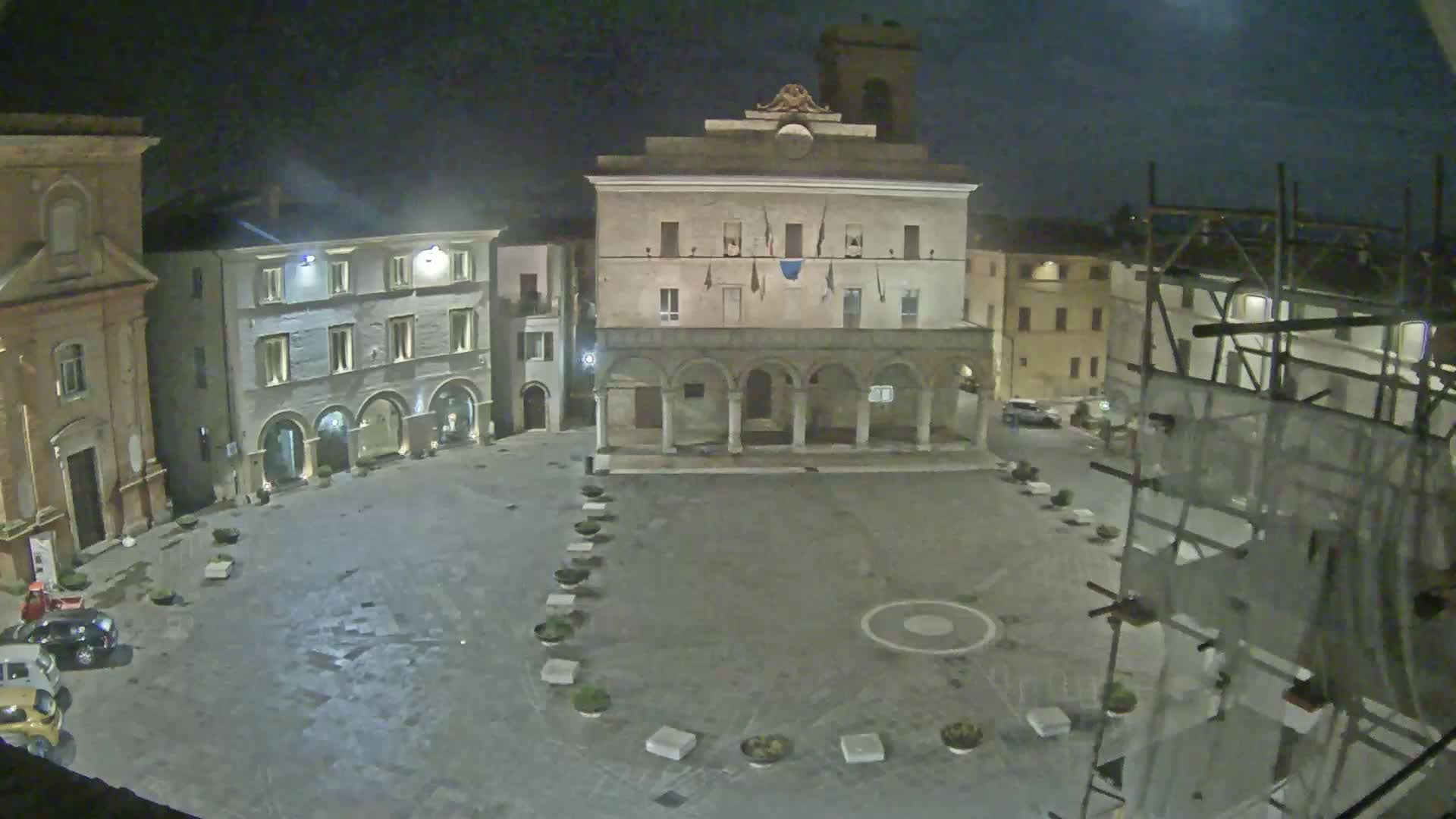 A nighttime view of a town square, featuring a large building with arched entryways, surrounded by other buildings under a dark, possibly overcast sky.