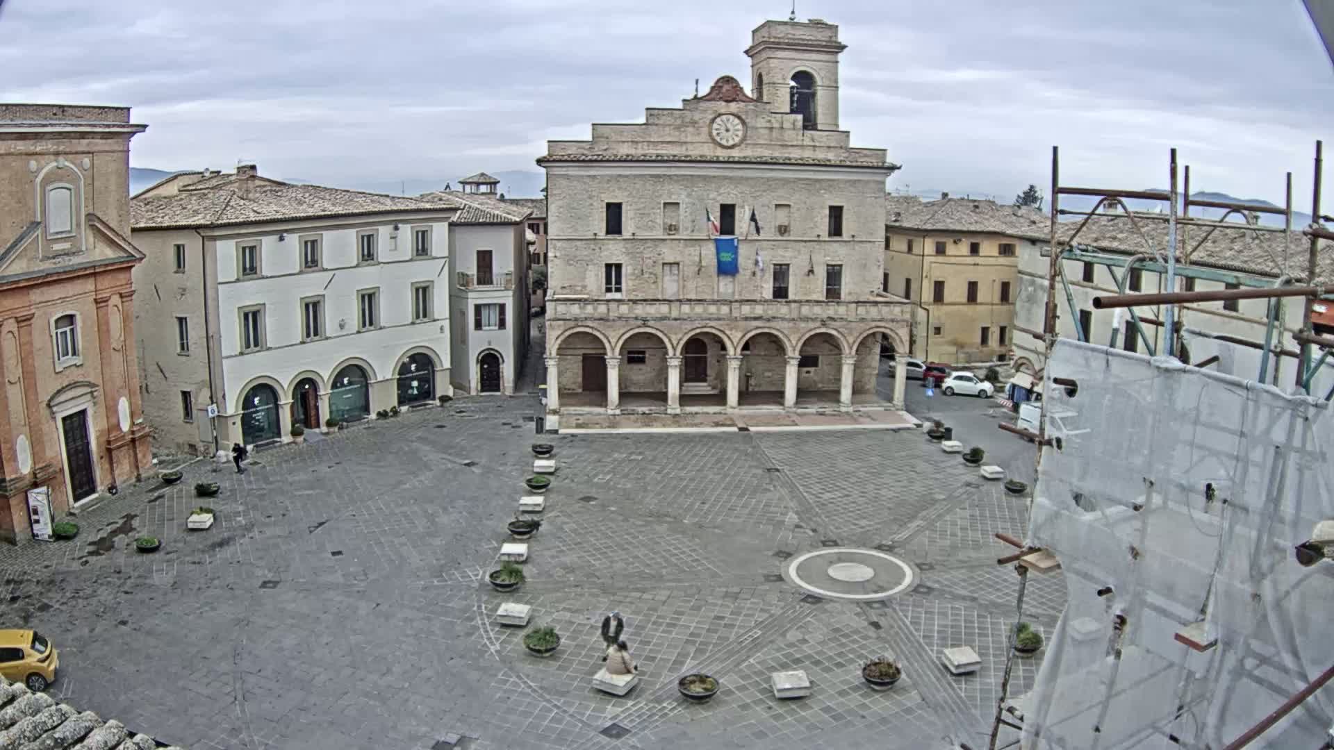 A large, paved town square is centered on a stone building with arched entryways, under an overcast sky.