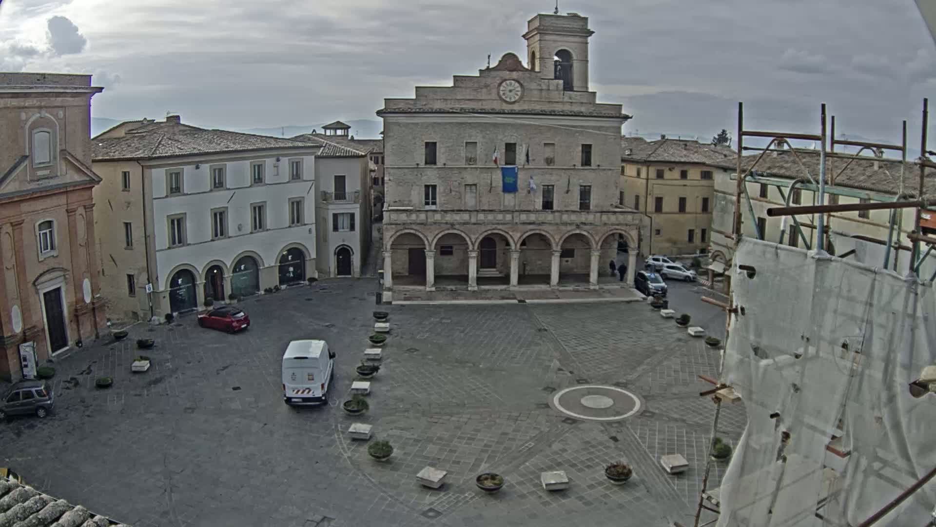A town square with several buildings, including a large building with a clock tower and arched entryway, under an overcast sky.