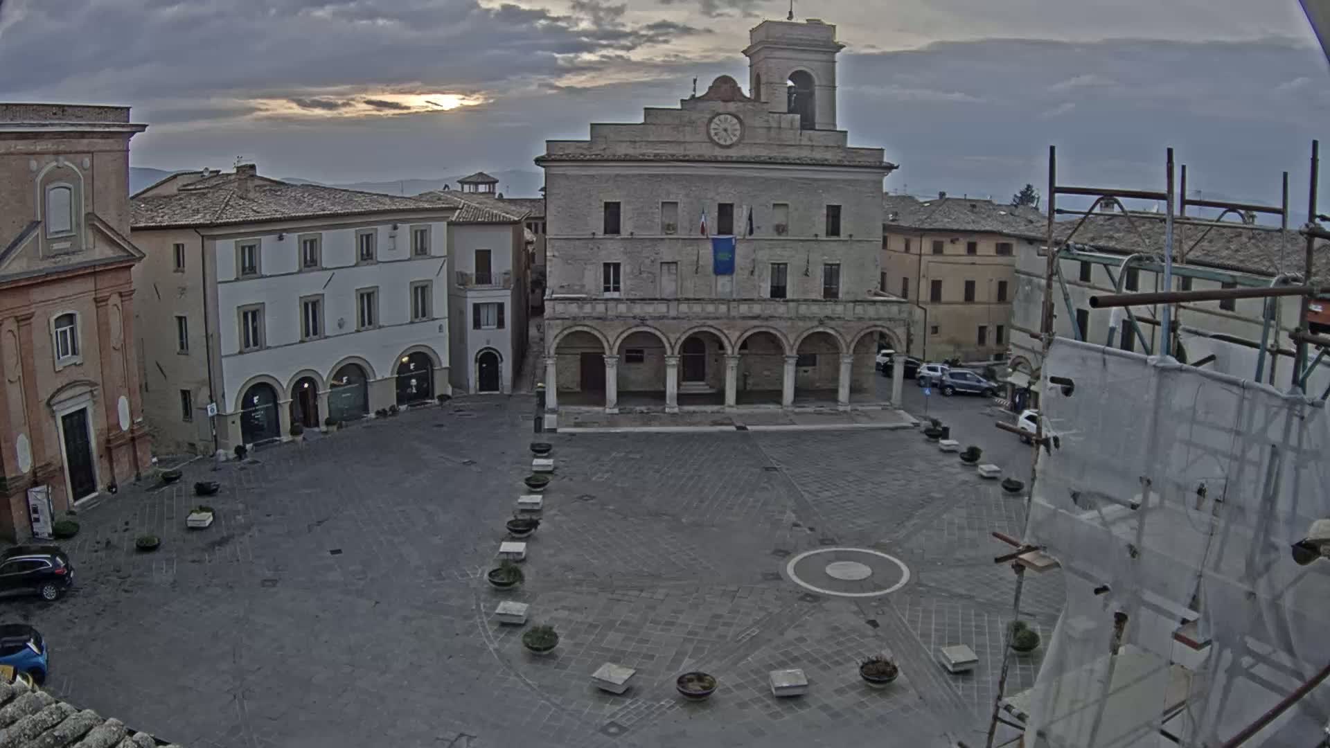 A large, empty town square with a clock tower and several stone buildings surrounding it under an overcast sky.
