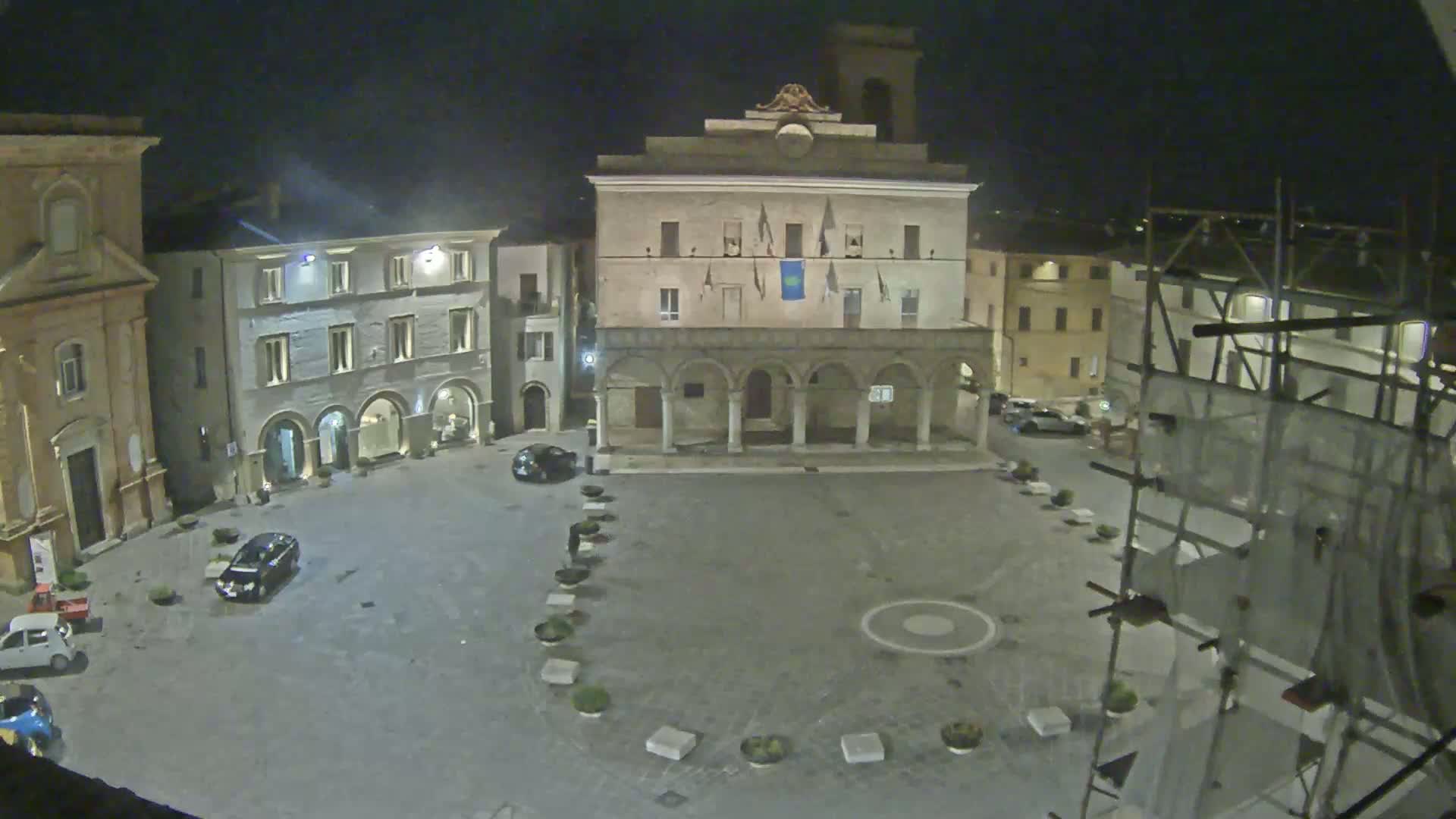 A nighttime view of a town square, with several buildings surrounding it, including a large building with arched entryways, under a dark sky.