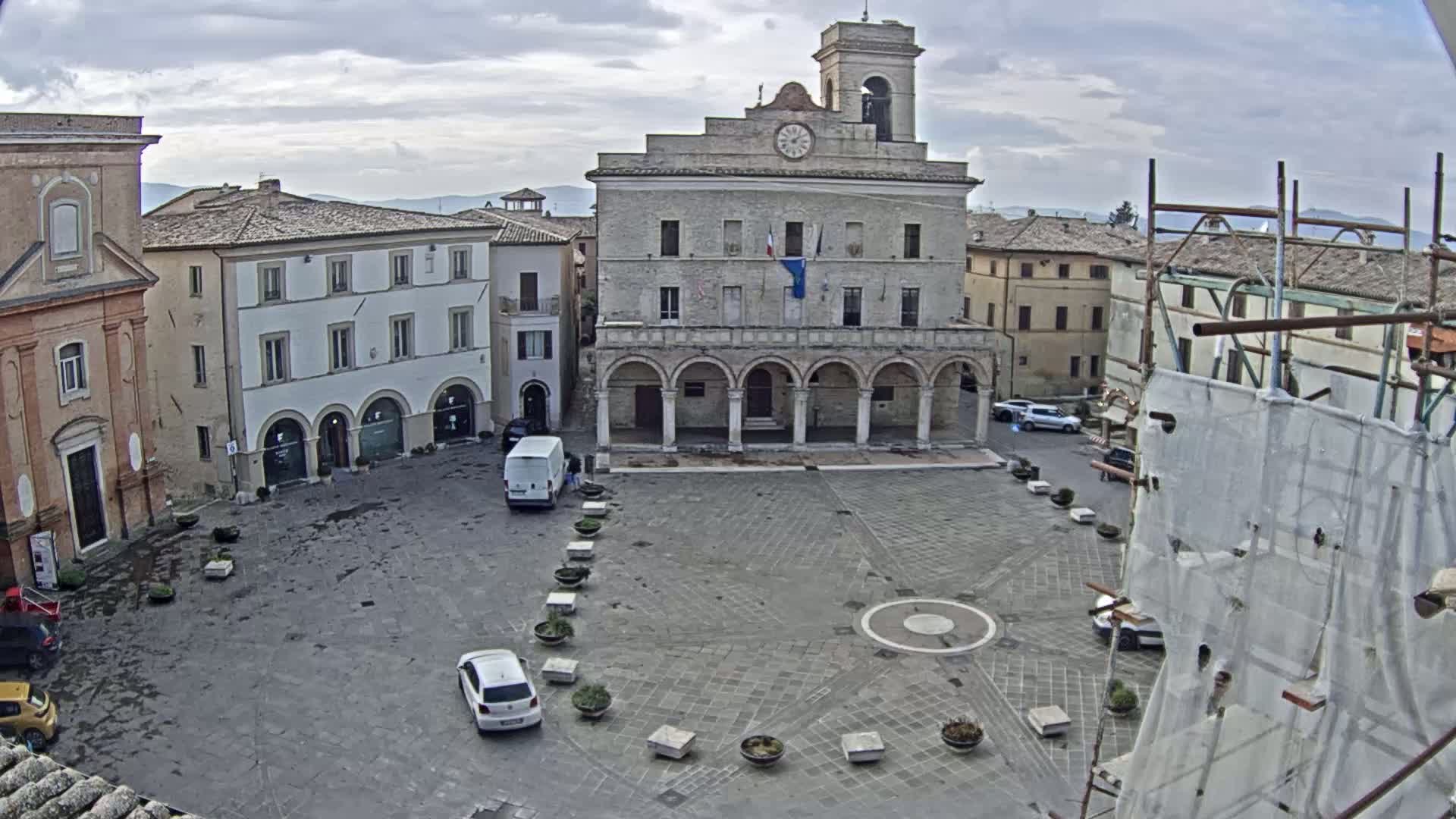 A town square with a large stone building, several smaller buildings, and a few cars under an overcast sky.