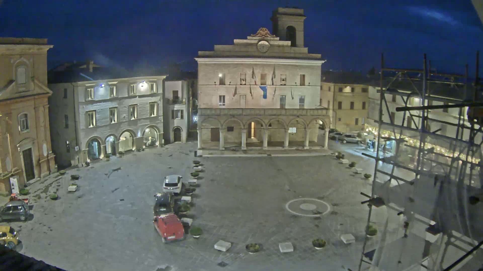 A town square at night, under a dark sky, is sparsely populated with a few cars parked along the side and a large building with arches in the center.