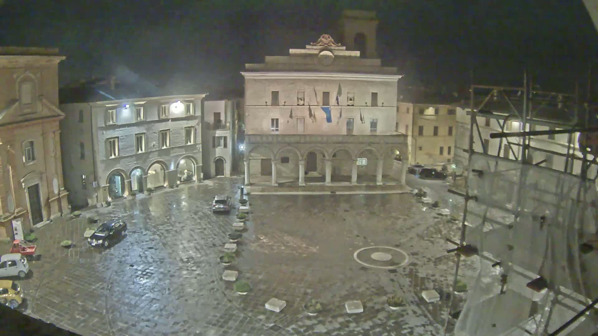 A rain-slicked town square at night is surrounded by historic buildings, one of which features a colonnade, under a dark, possibly stormy sky.