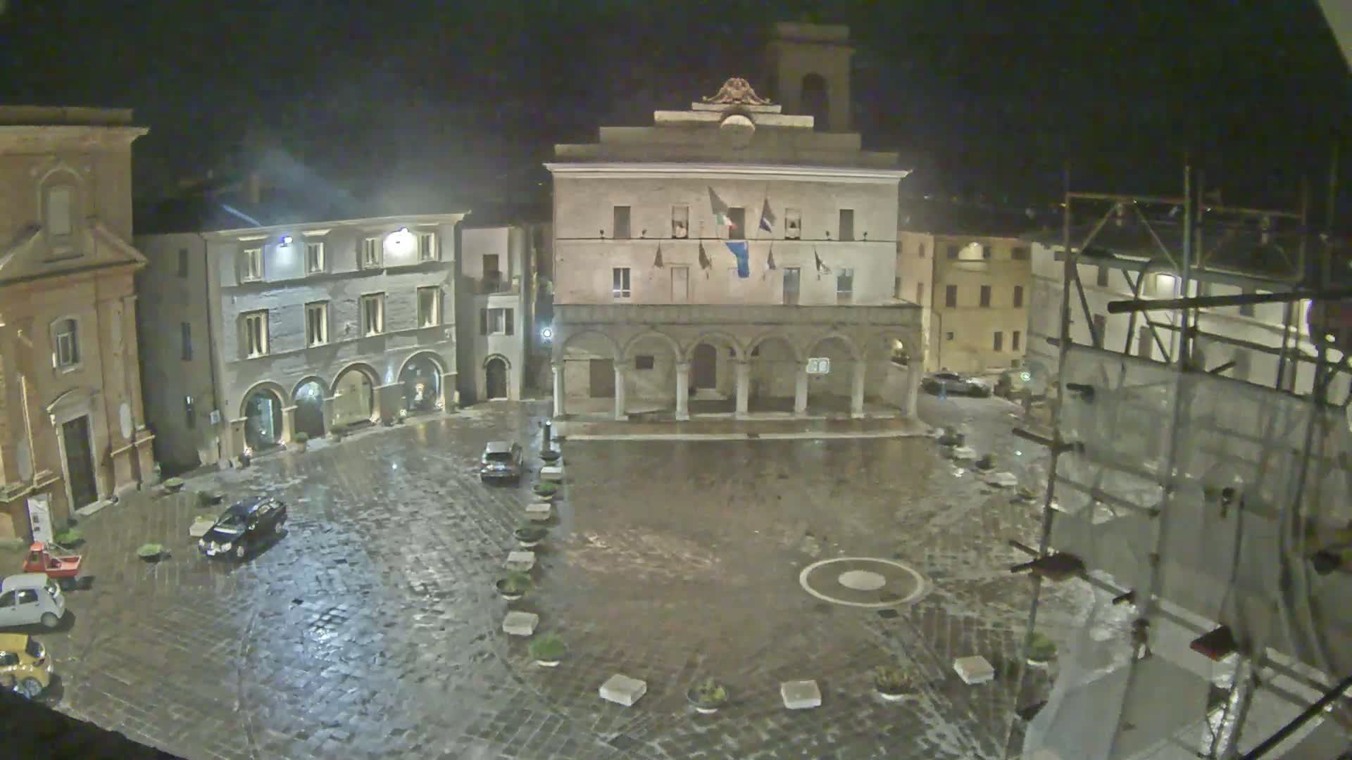 A rain-slicked town square at night, surrounded by historic buildings, one of which is a large structure with arched colonnades.