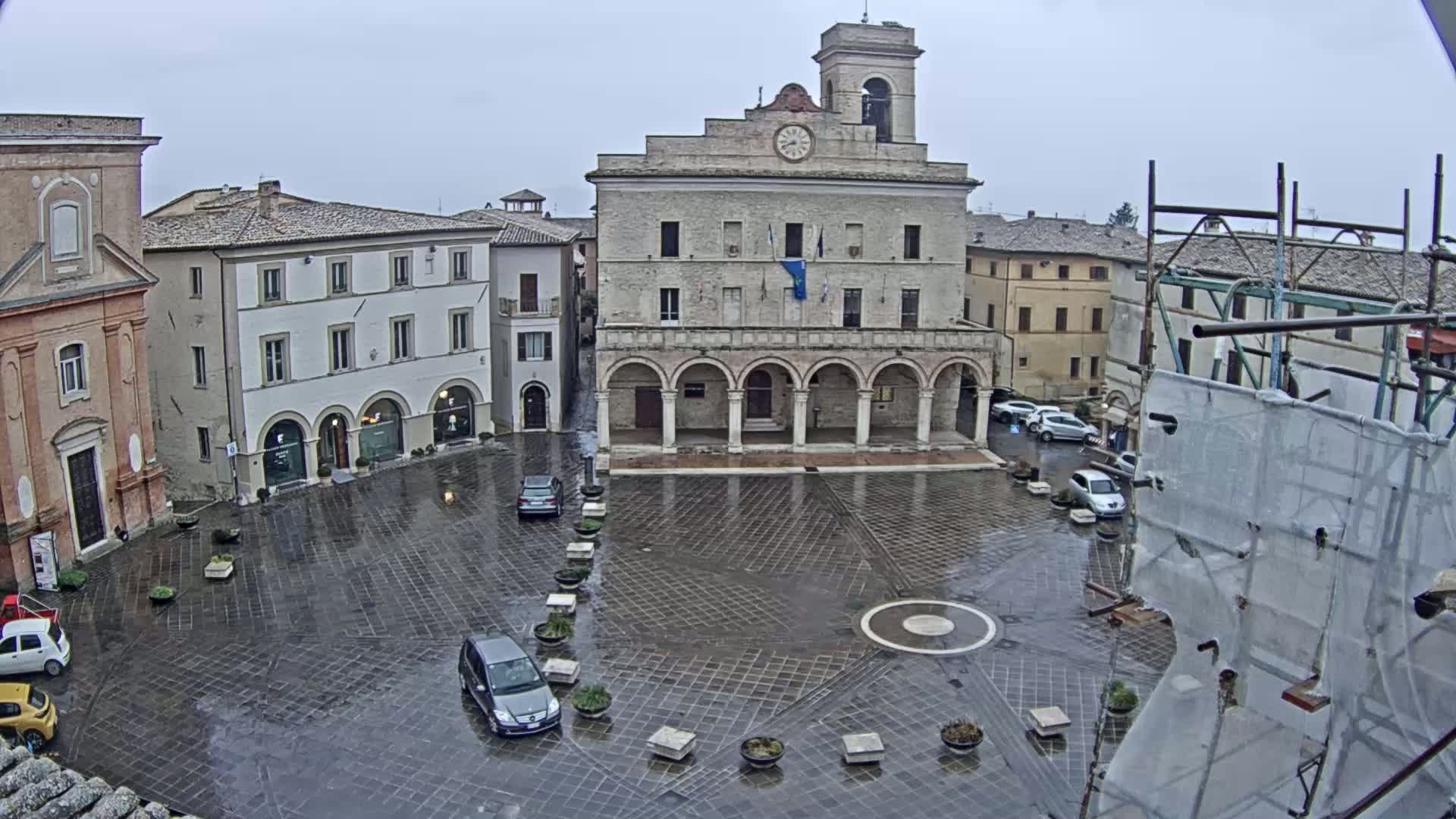 A wet, paved town square is centered on a large stone building with arched entryways, surrounded by other buildings, a few cars, and scaffolding.
