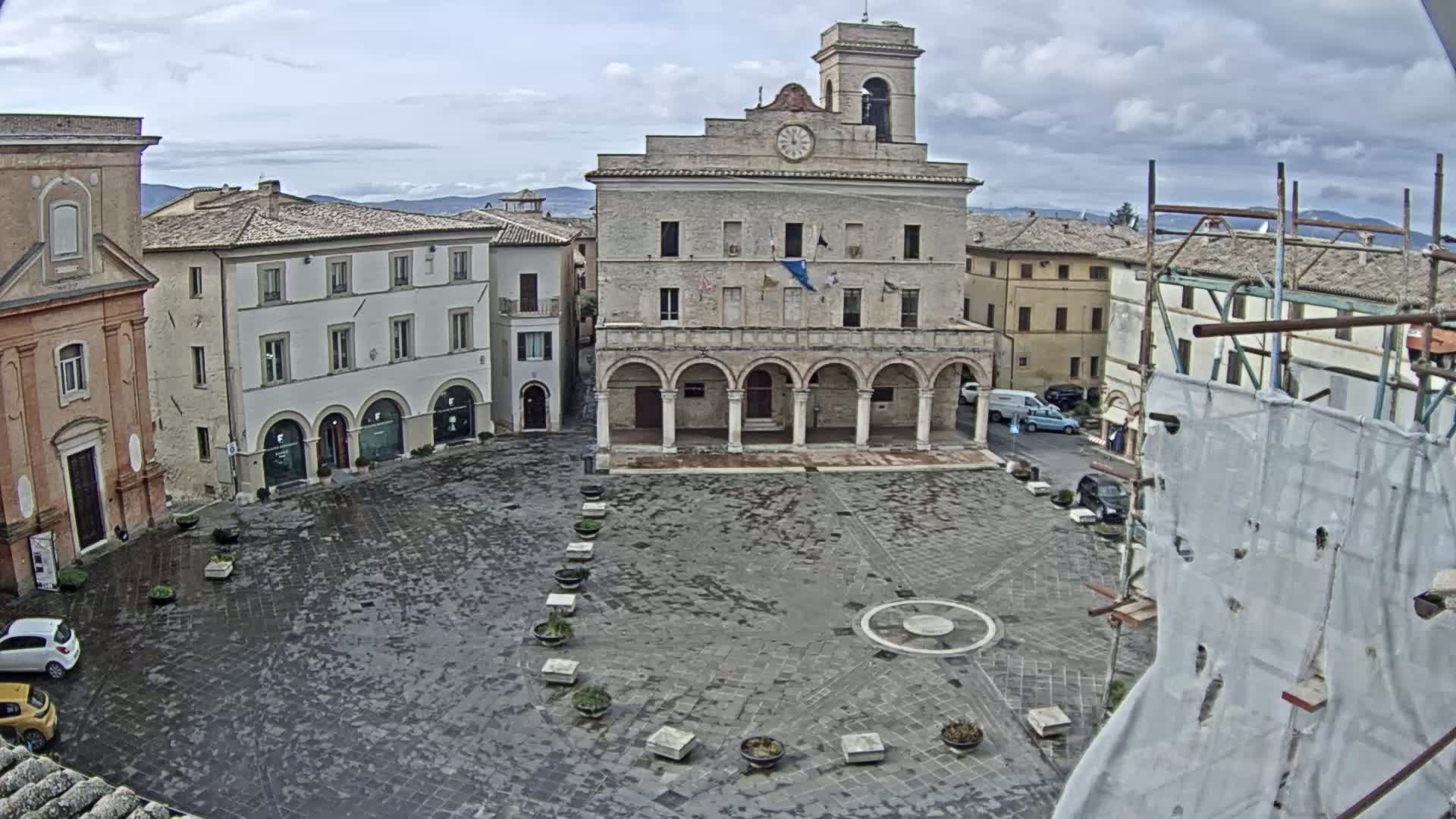 A large, stone plaza with a prominent building featuring arched entryways and a clock tower is viewed on an overcast day.