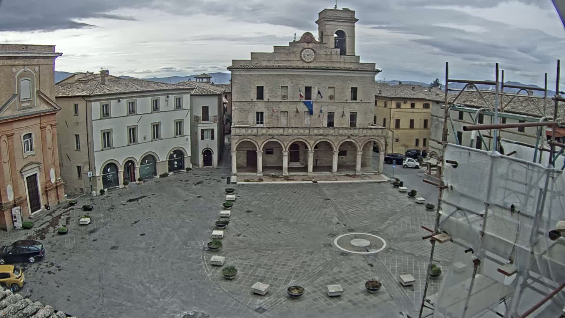 A town square with a large stone building, featuring arched colonnades and a clock tower, under a cloudy sky.