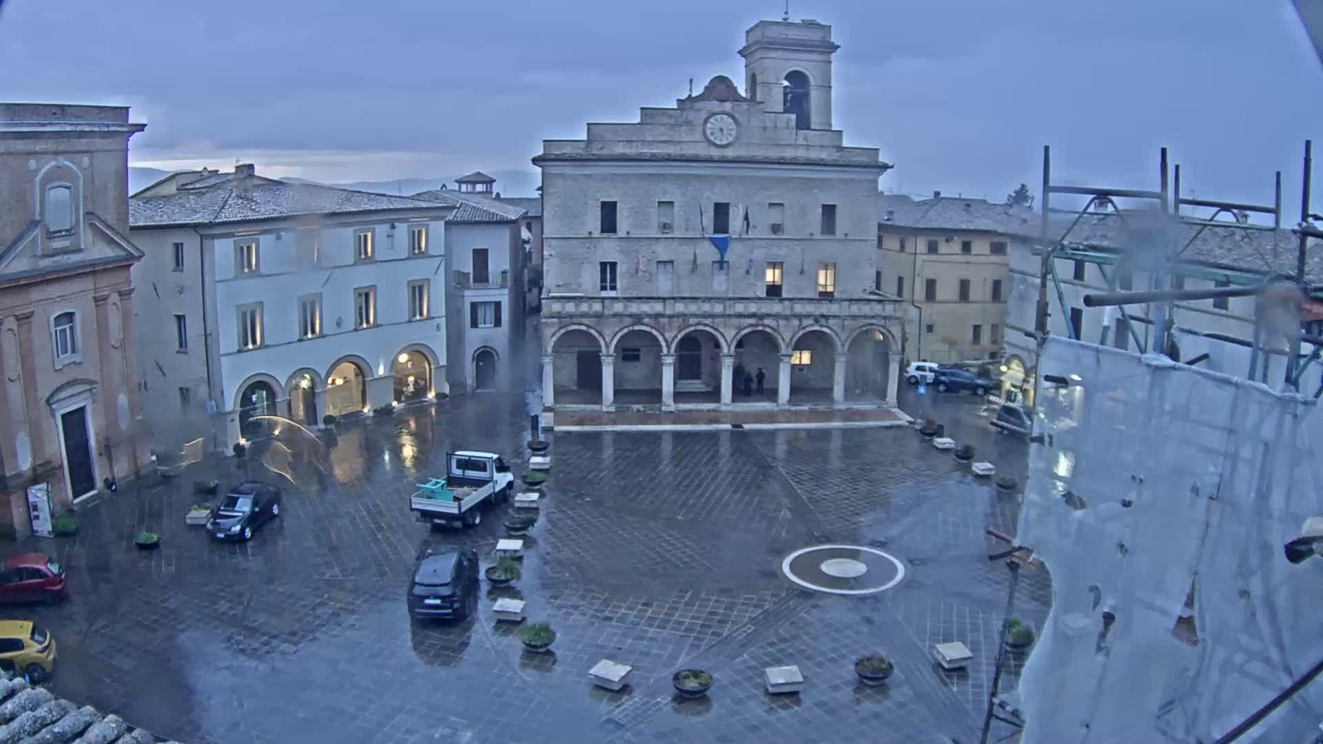 A rain-soaked town square is centered on a large stone building with arched entryways, surrounded by other buildings under an overcast sky.
