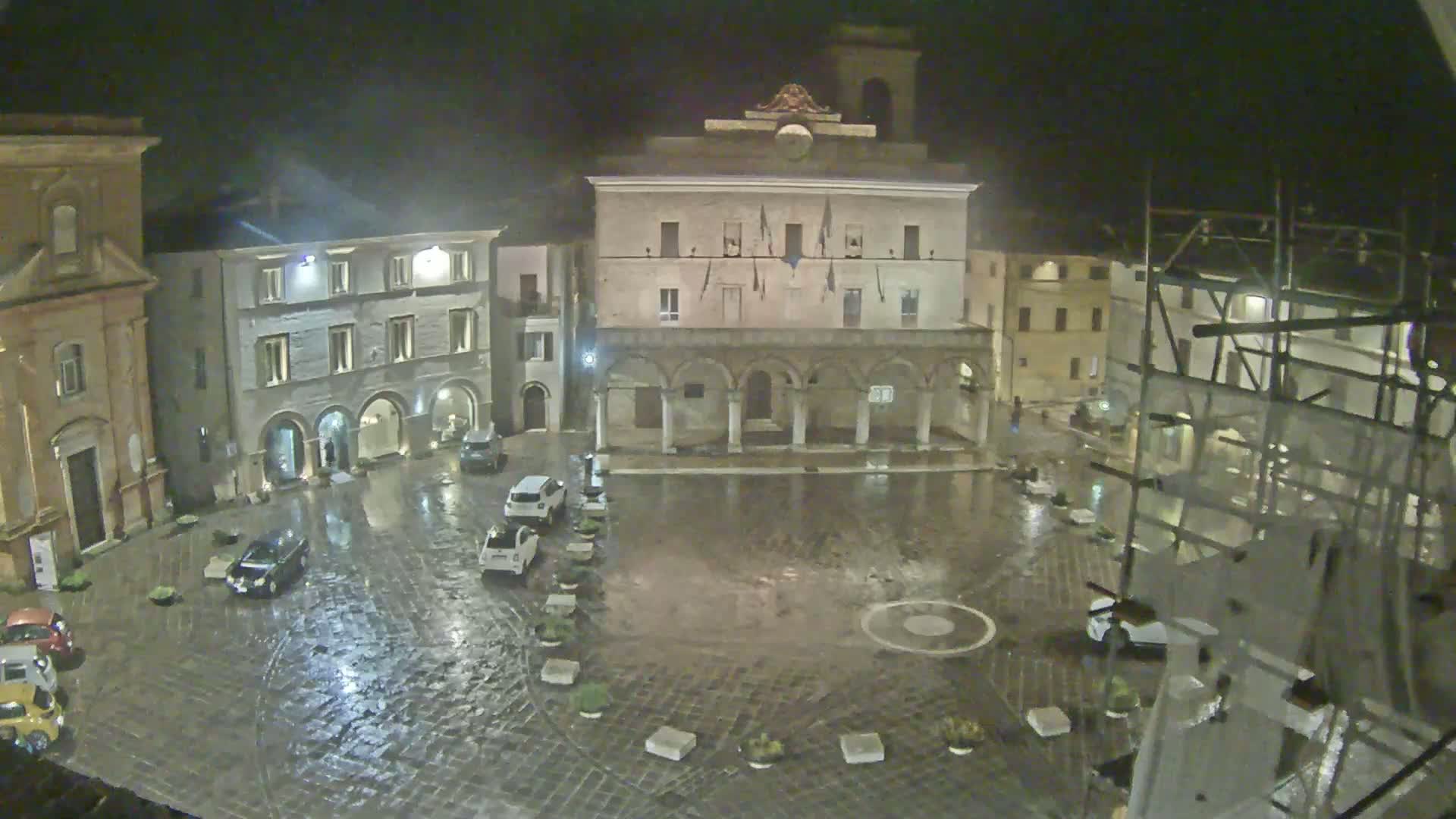 A rain-soaked town square at night, surrounded by historic buildings, with a few cars parked.