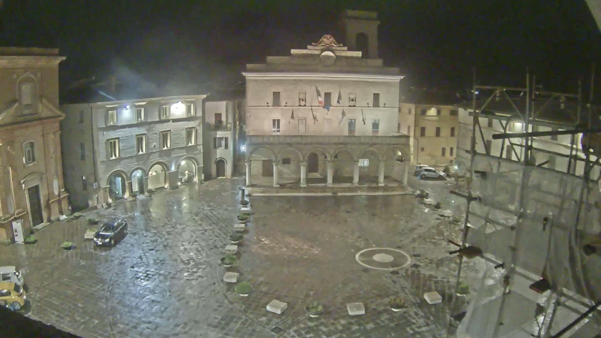 A rain-slicked town square at night, surrounded by historic buildings, one of which has a colonnaded facade, with scaffolding visible on one side.