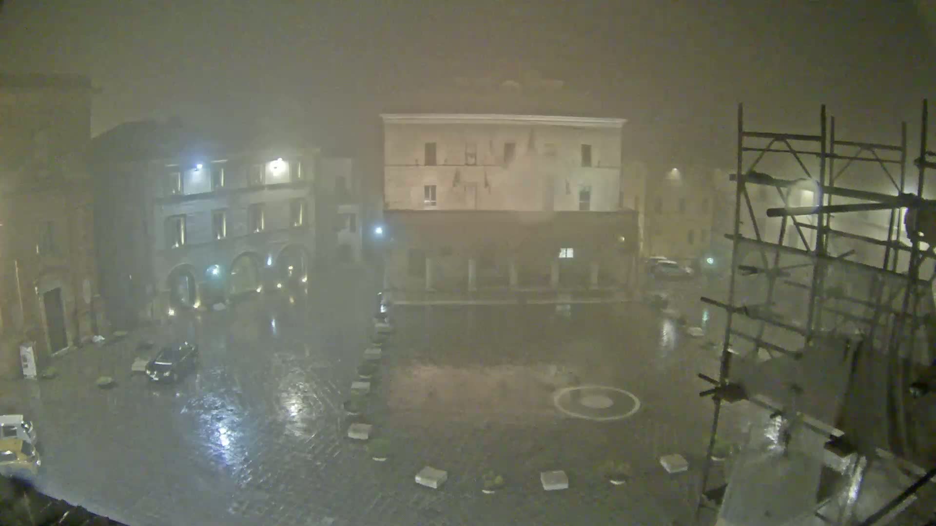 A rain-soaked town square at night is seen under a heavy fog, with several buildings surrounding it and scaffolding to the right.