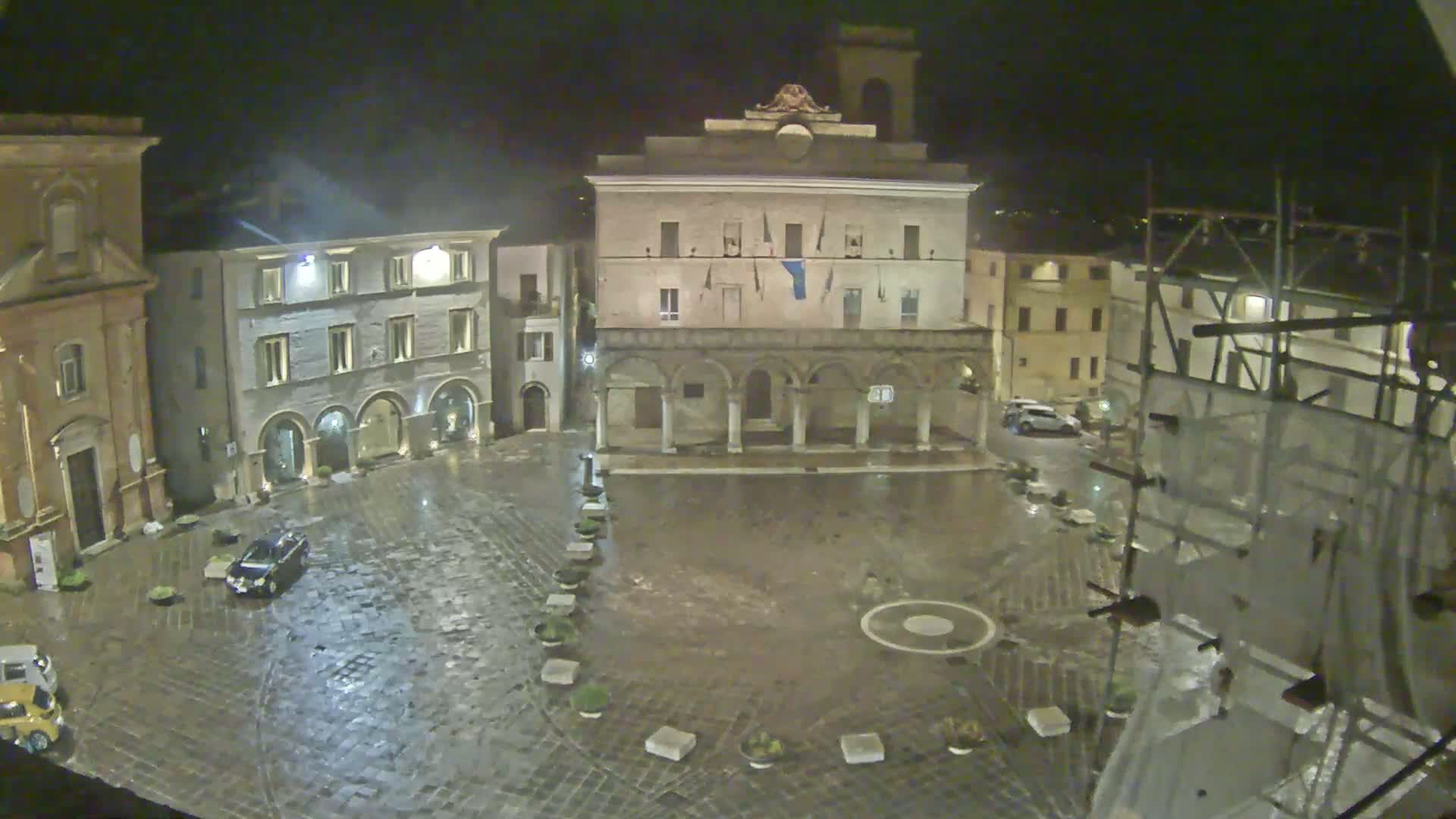 A rain-slicked, cobblestone town square at night is surrounded by historic buildings, one of which has scaffolding on it.