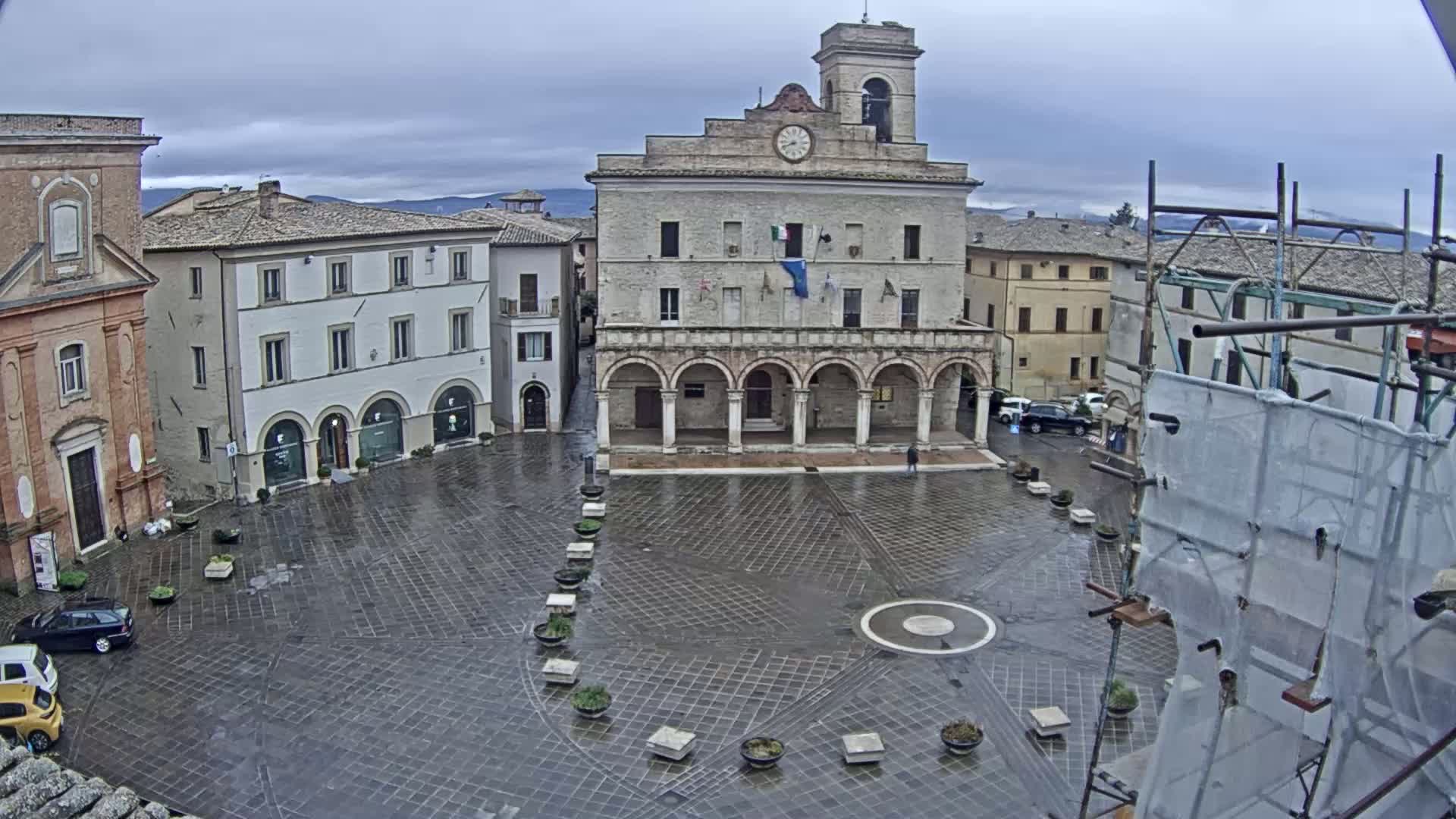A wet, paved town square is centered on a large, light-colored building with arched entries, surrounded by other buildings under a cloudy sky.