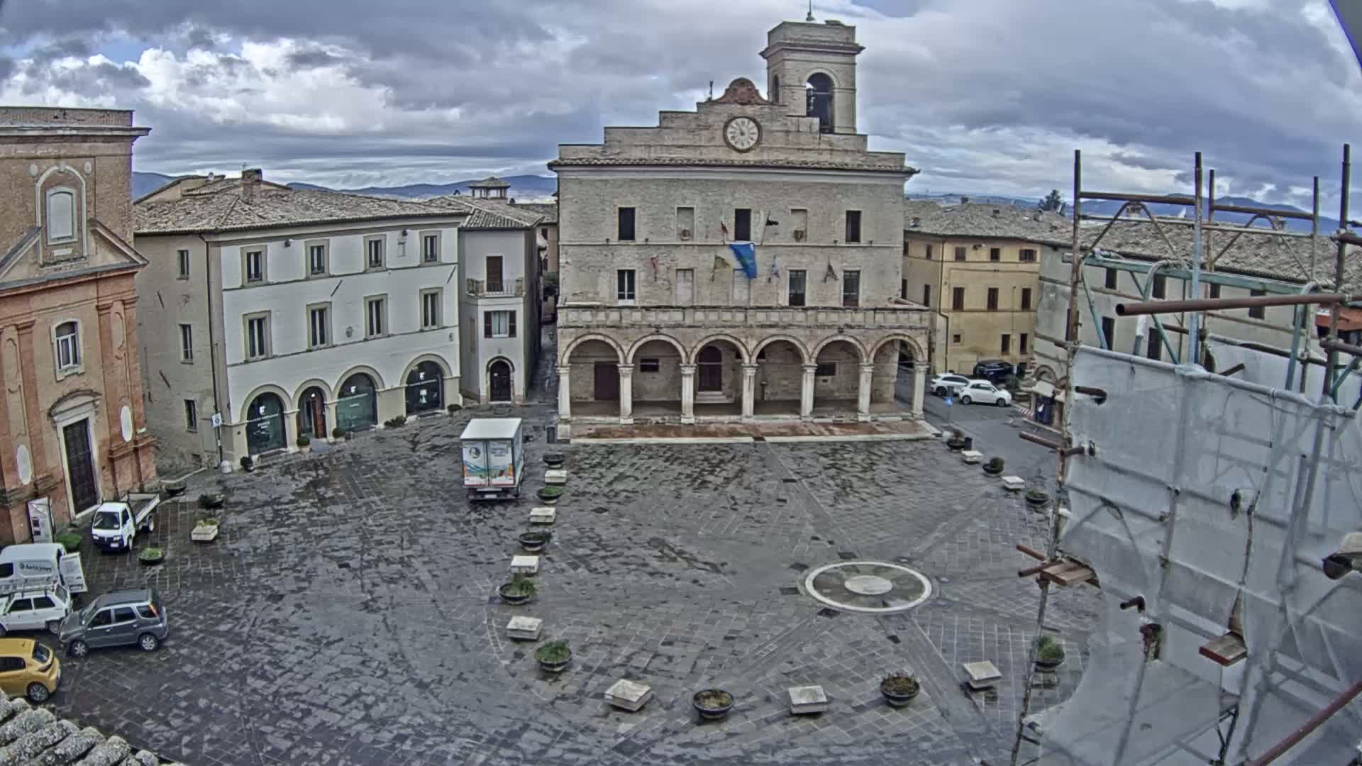 A large, stone plaza is shown under a cloudy sky, centered on a large building with arched entryways and a clock tower.