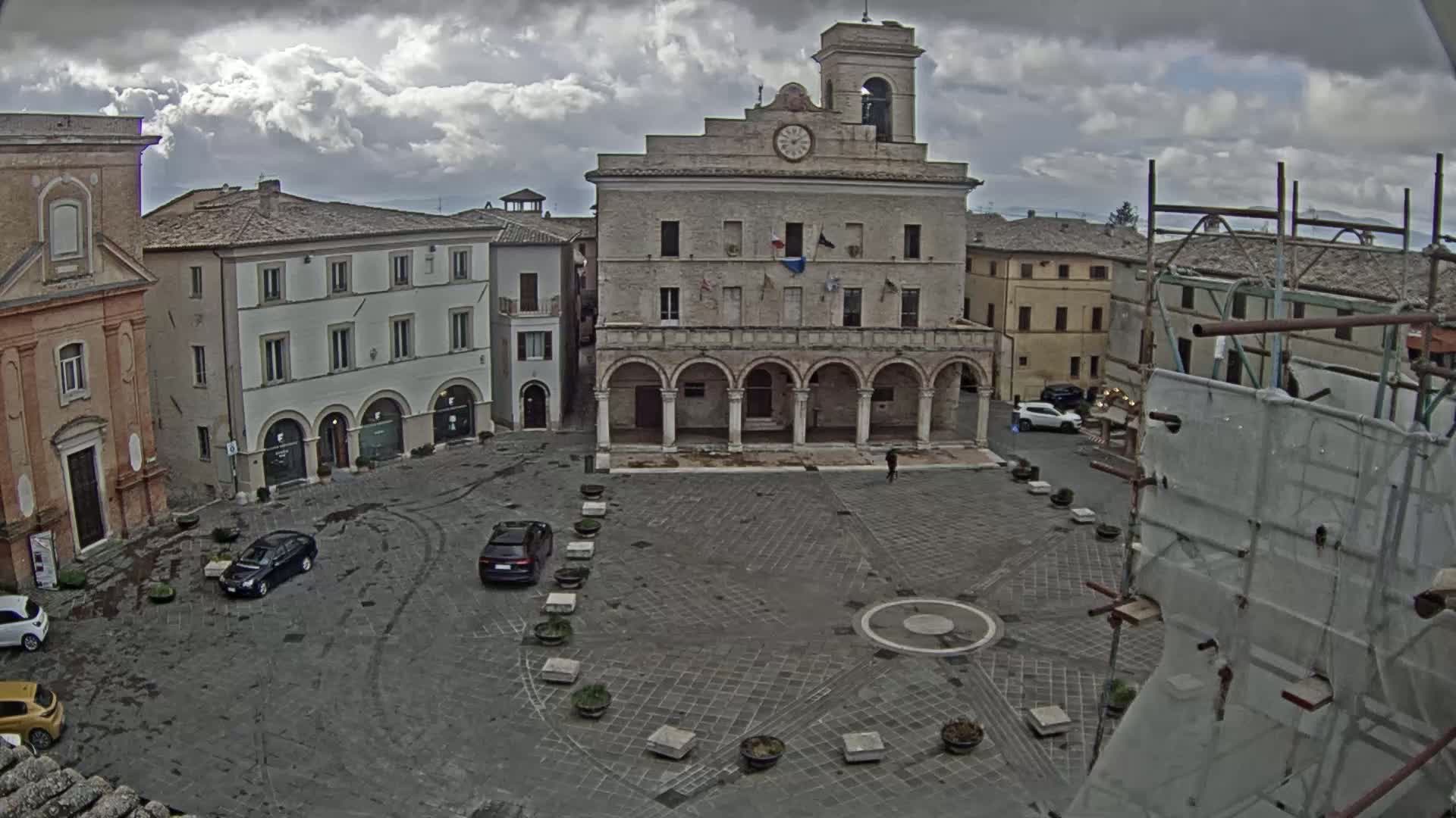 A town square with several parked cars, older buildings, and a clock tower under a cloudy sky.
