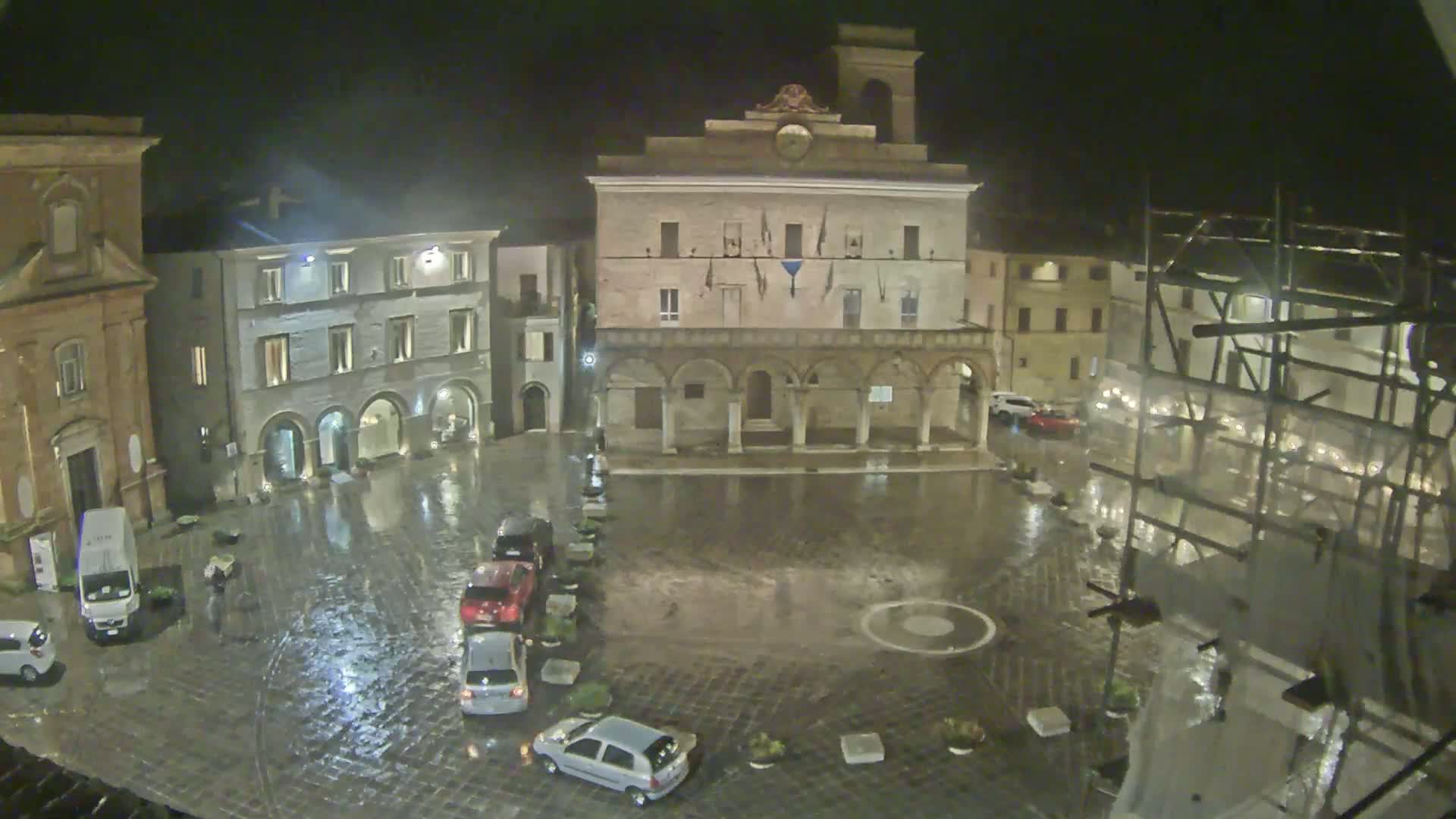 A rain-slicked town square at night, surrounded by buildings, with several cars parked along the edge.