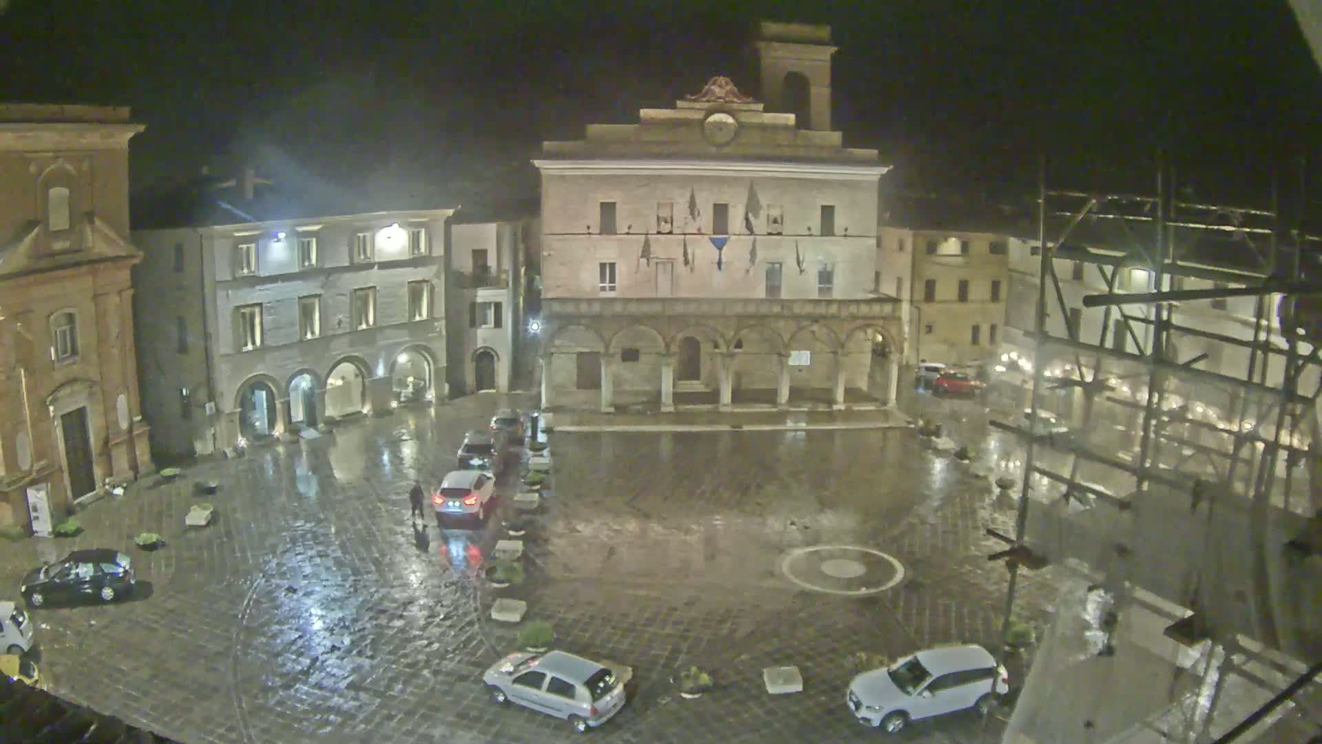 A rain-soaked town square at night, surrounded by historic buildings, with a few cars parked and one person walking.