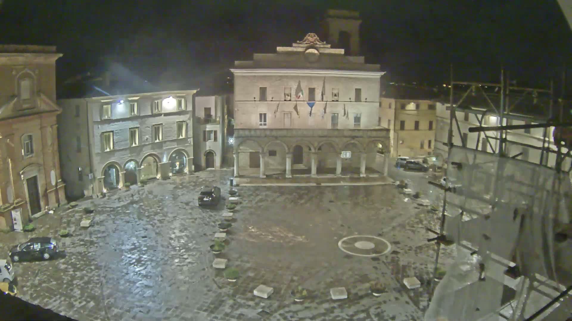 A wet, cobblestone town square at night is surrounded by historic buildings, one of which is a large building with columns and flags, and scaffolding is visible on a building to the right.