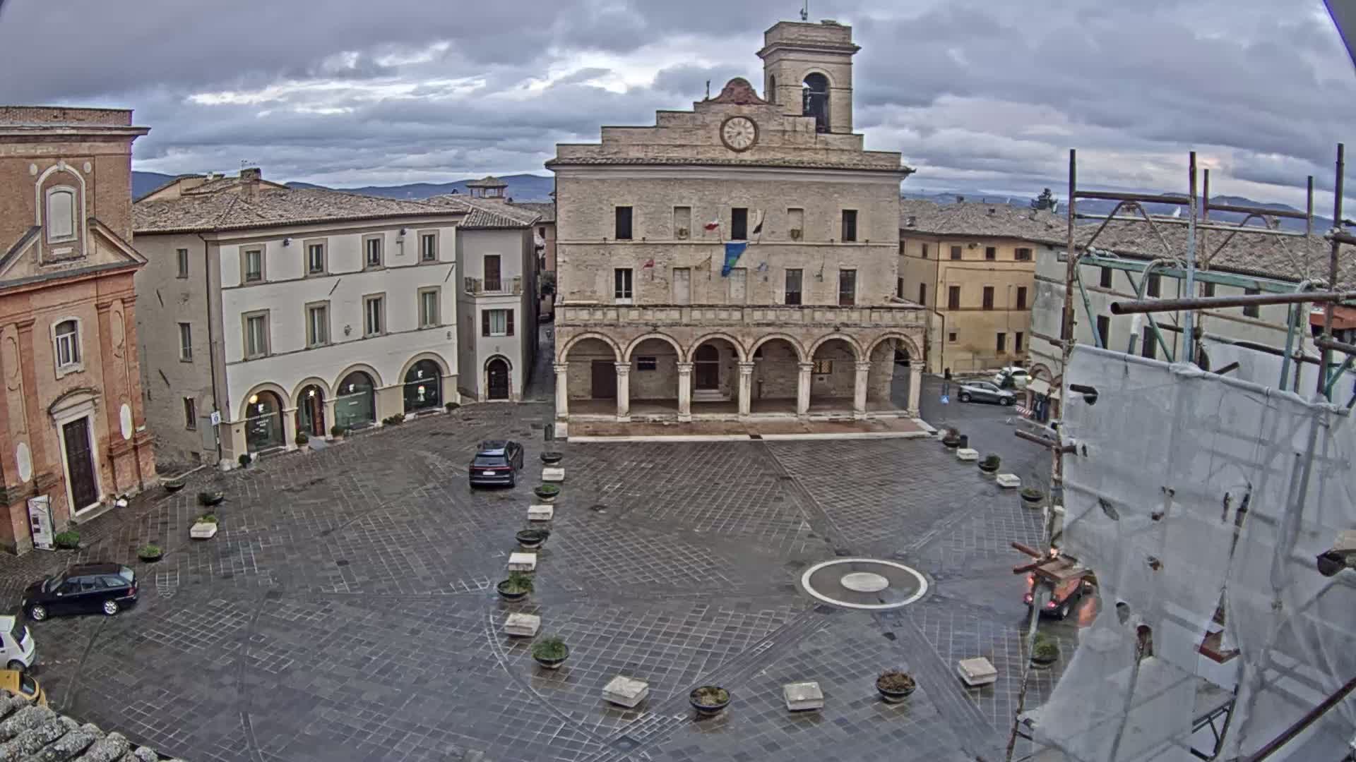 An overcast day reveals a town square with a large, light-colored building featuring arched entryways, surrounded by other buildings, some with scaffolding, and a few cars parked in the square.