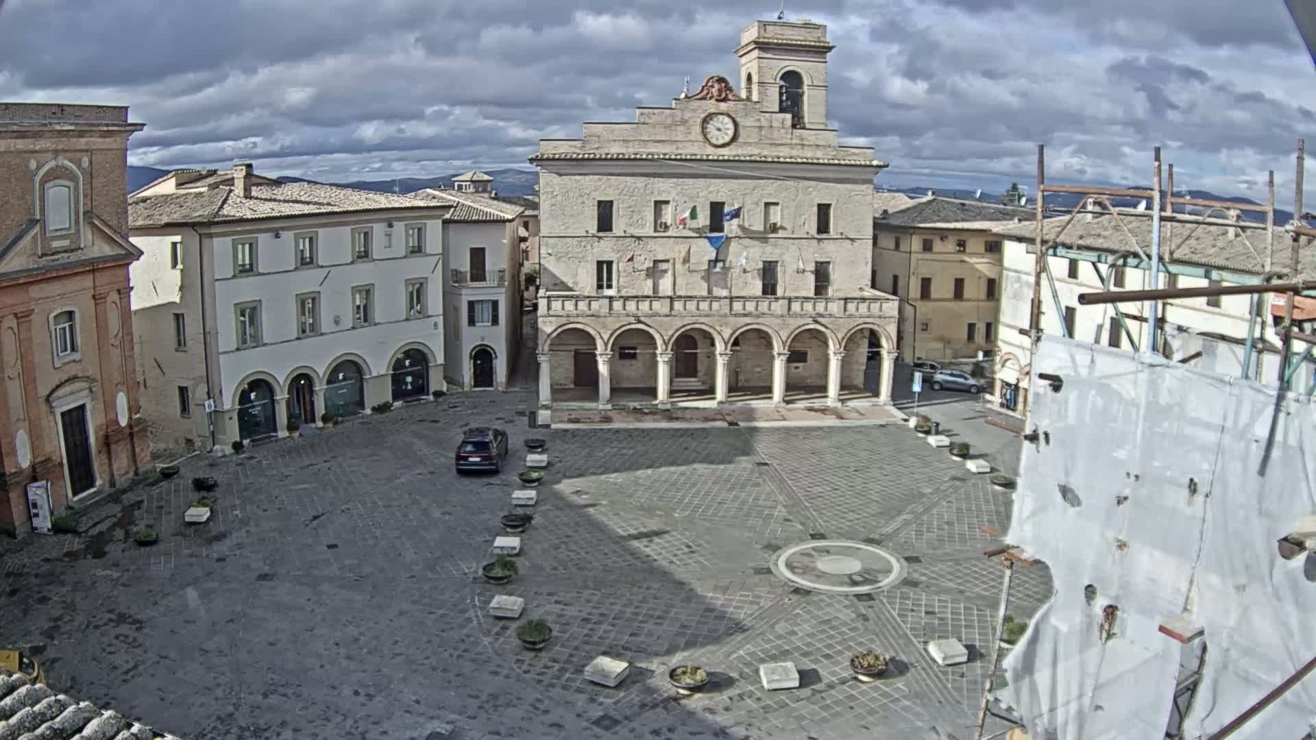 A large, stone plaza with a prominent town hall building is shown under a cloudy sky, surrounded by other buildings.