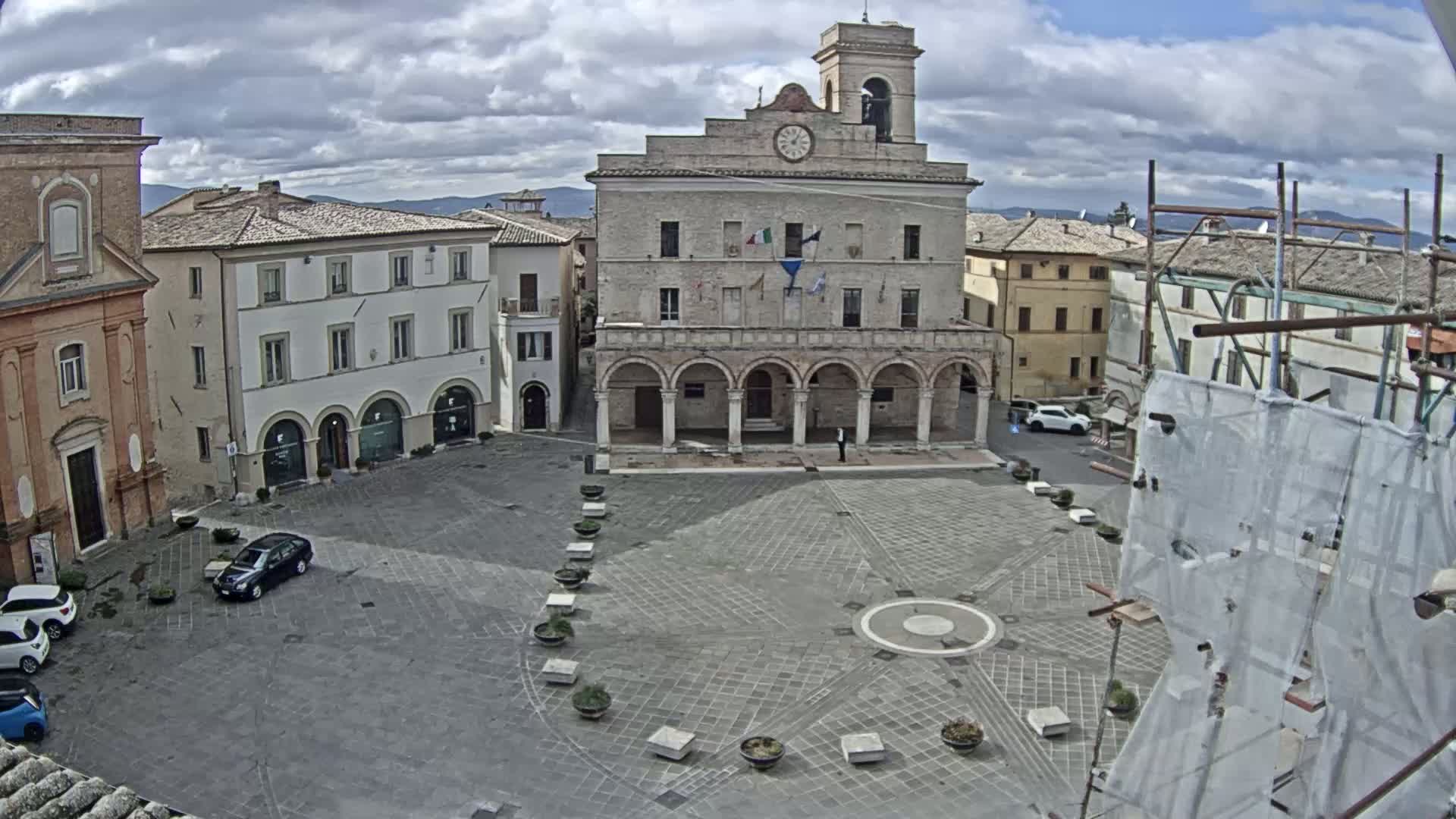 A town square with a large stone building featuring arched columns and a clock tower is partially surrounded by other buildings under a cloudy sky.