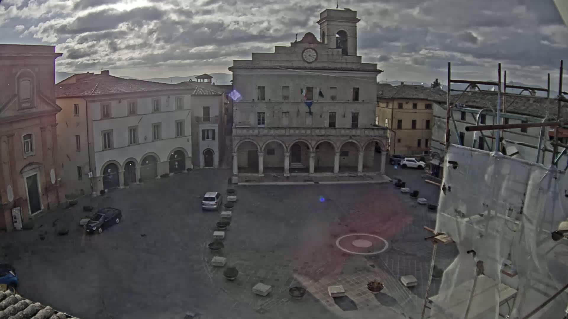 A town square with a large building featuring arched walkways, a clock tower, and several cars parked in the square, under a cloudy sky.