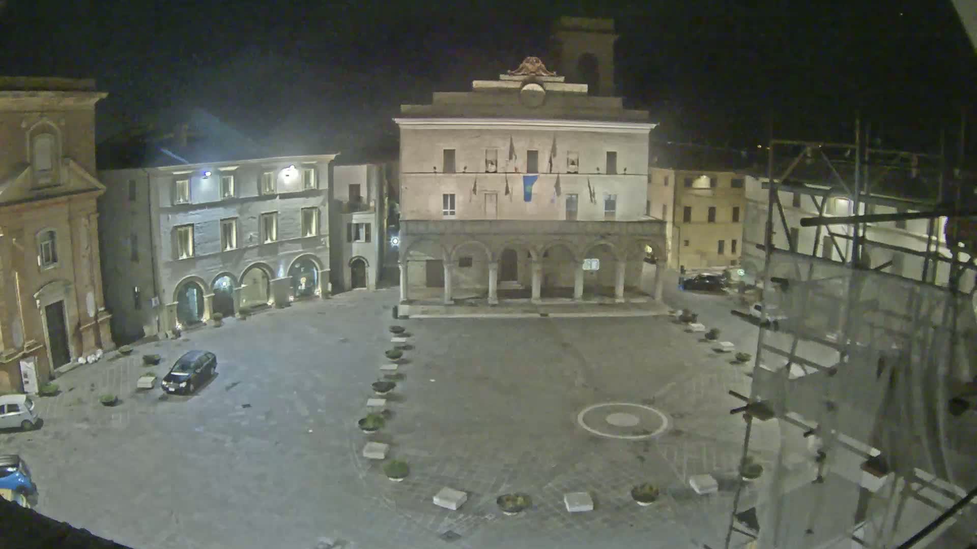 A nighttime view of an empty town square, centered on a large building with arches, surrounded by other buildings under a clear, dark sky.