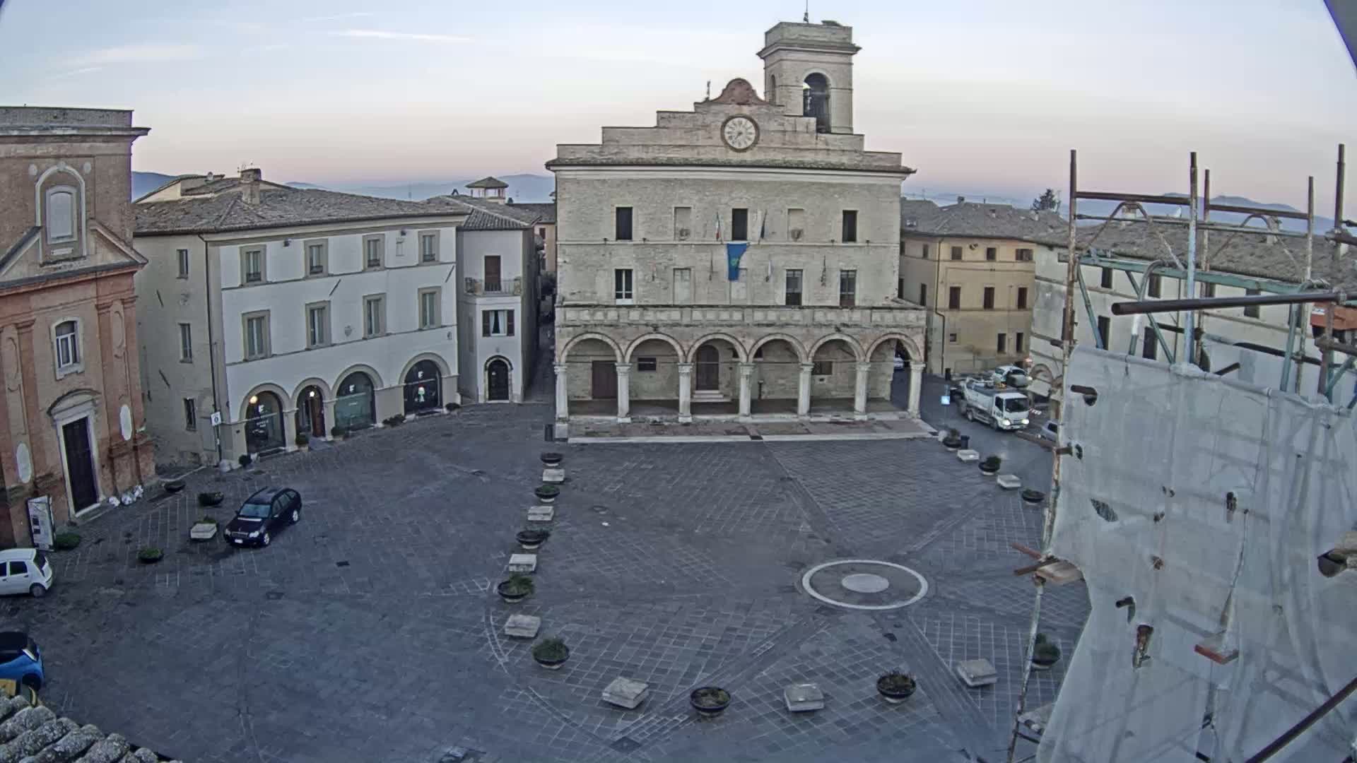 A town square, under a pale dawn sky, features a large, pale stone building with arched entryways and a clock tower, surrounded by other buildings and a few parked cars.