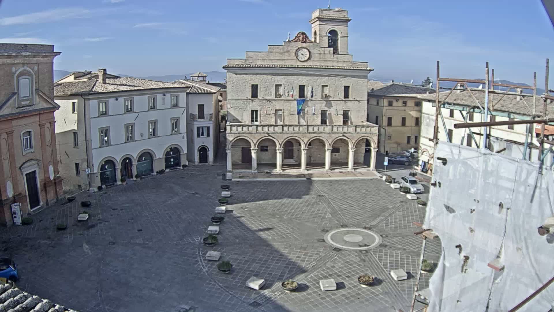 A large, paved town square is surrounded by buildings, including a prominent stone structure with arches, under a clear, sunny sky.