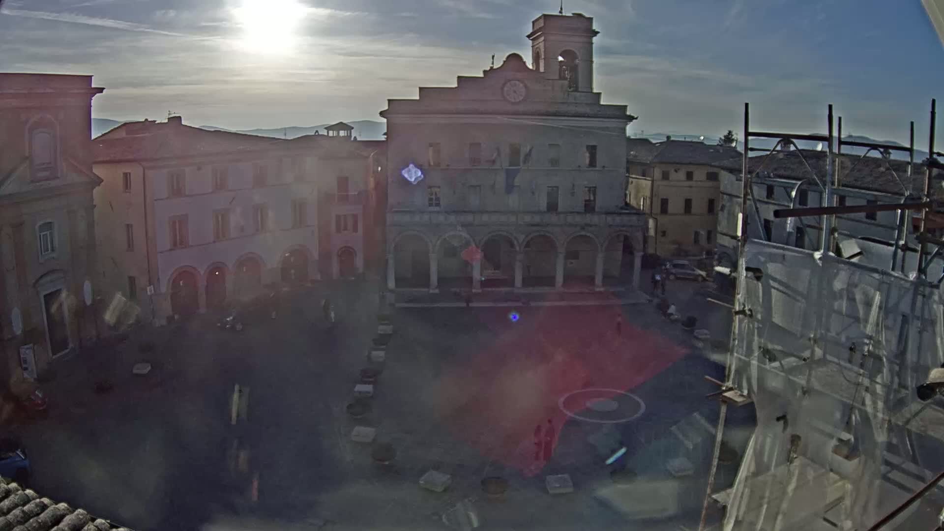 A sunlit town square with several buildings, including a large stone structure with arched entrances and a clock tower, under a mostly clear sky.