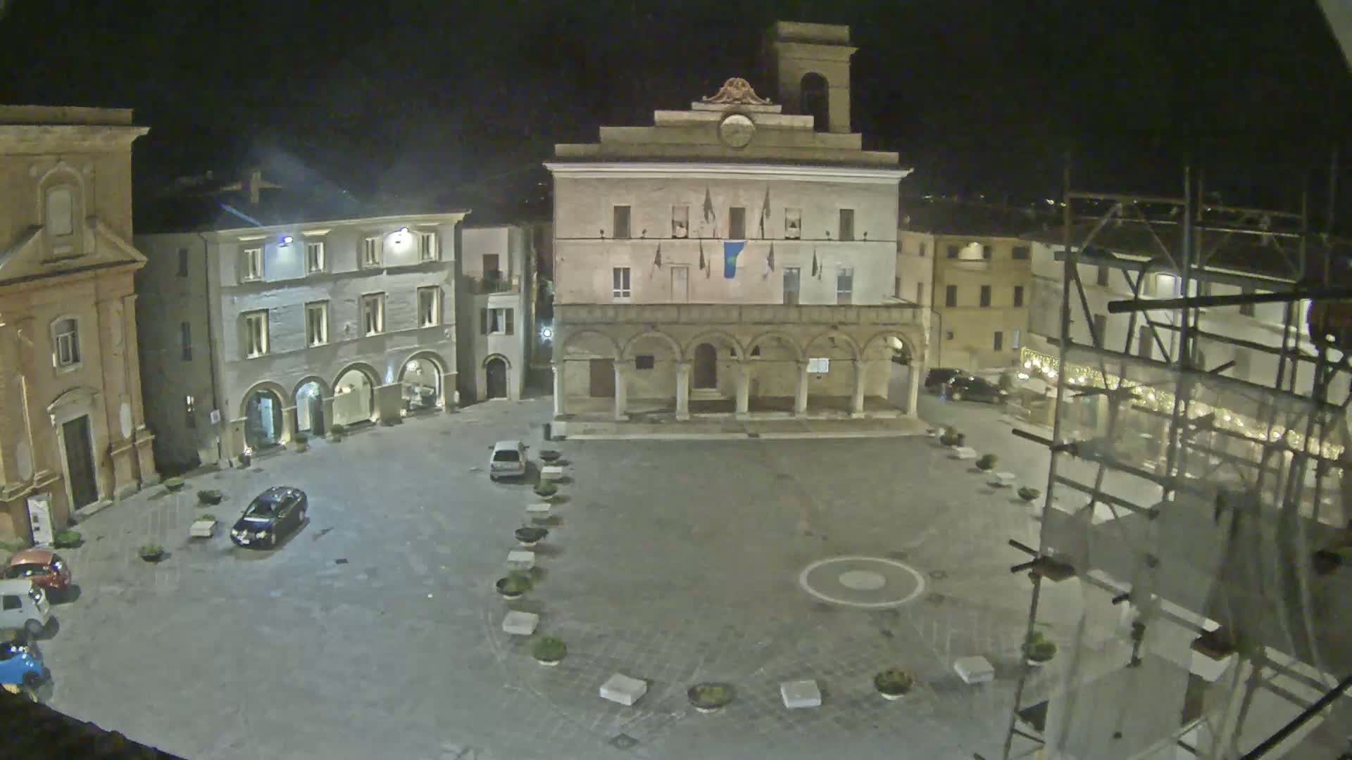 A nighttime view of an empty town square, featuring several buildings and a few parked cars under dark skies.
